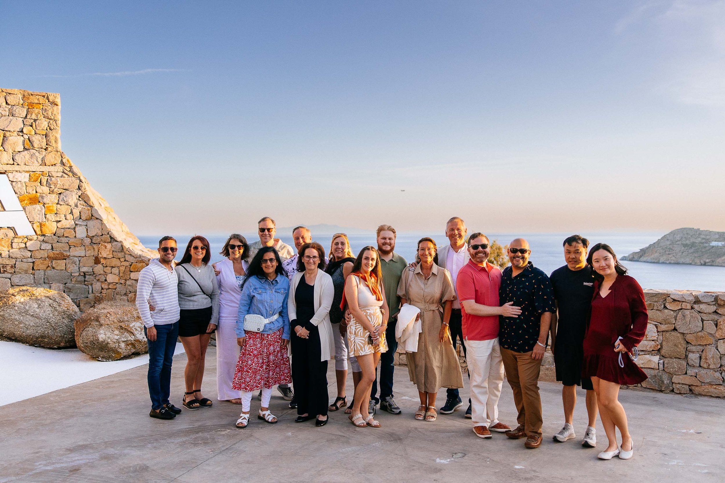 Group of 17 people posing outdoors on a stone platform with ocean and land in the background during sunset.