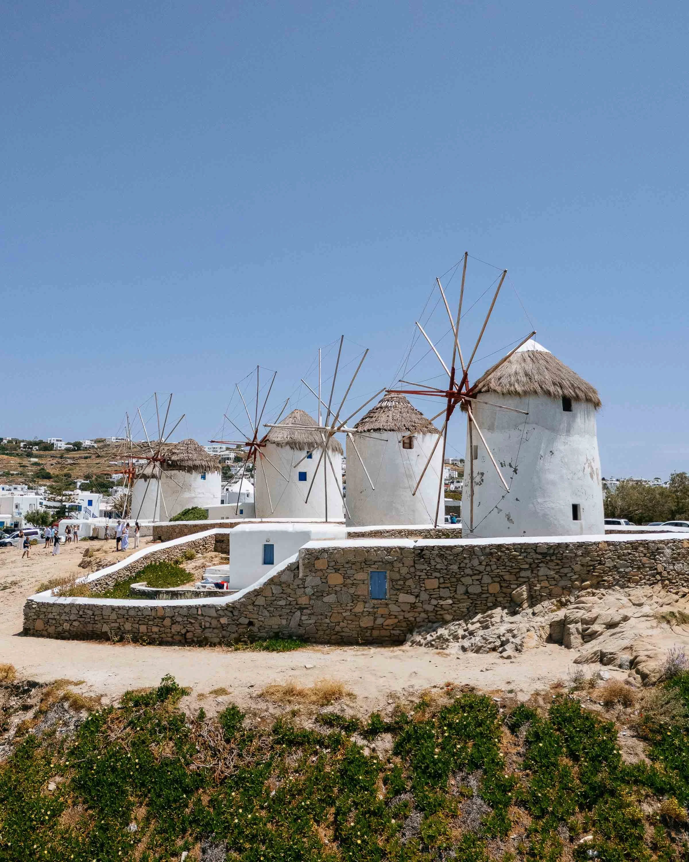 Three white traditional windmills with thatched roofs on a stone platform against a clear blue sky, with a distant landscape and small buildings in the background.