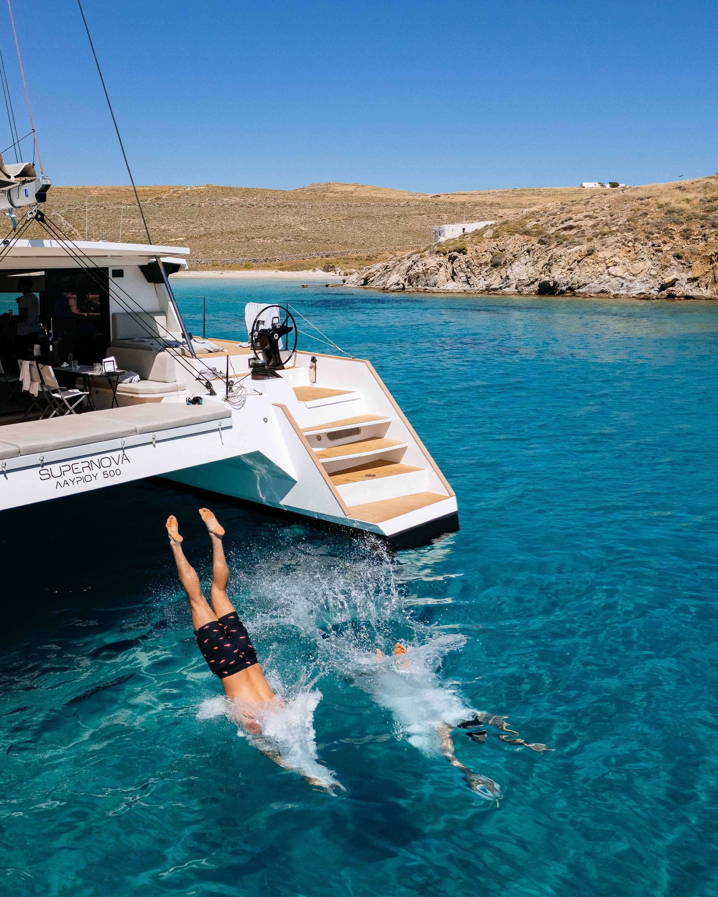 Person diving into the water next to a white yacht with stairs, on a sunny day with hilly landscape in the background.