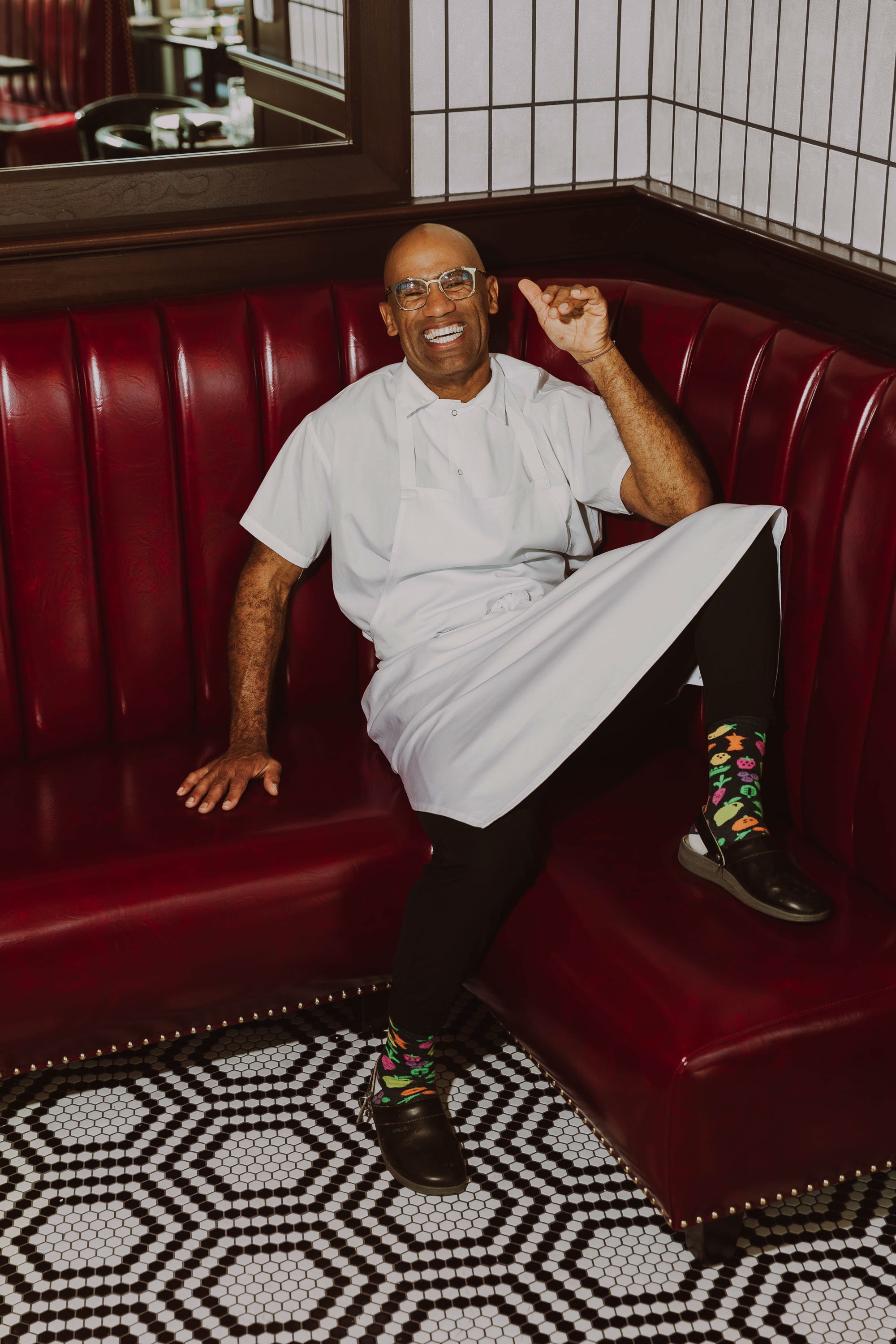 A man wearing glasses, a white shirt, and colorful socks is sitting on a red leather booth in a restaurant. He is smiling and appears to be laughing. The restaurant has black and white tile flooring and a mirror on the wall behind him.