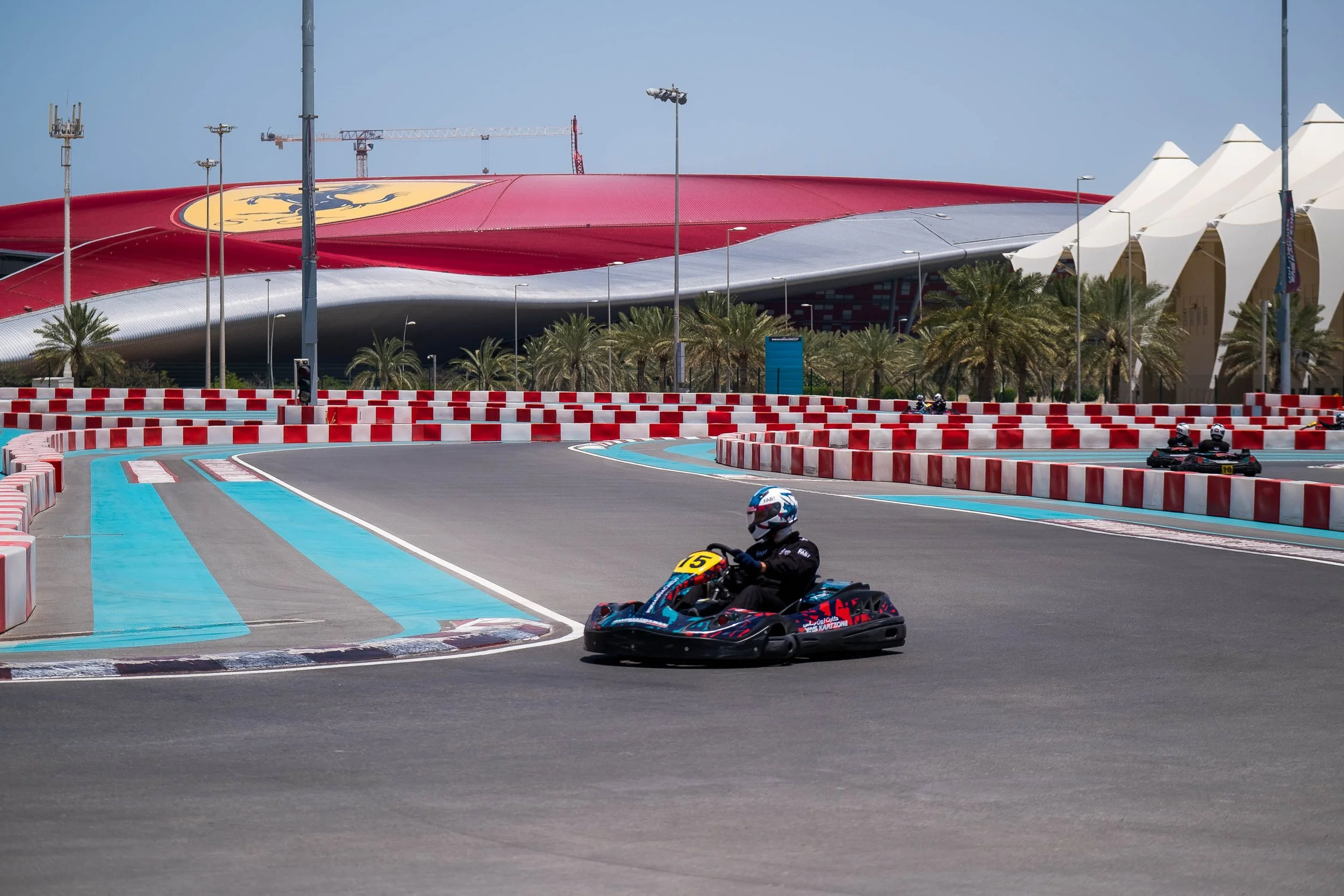 A person driving a go-kart on an outdoor race track with a large stadium building, palm trees, and blue sky in the background.