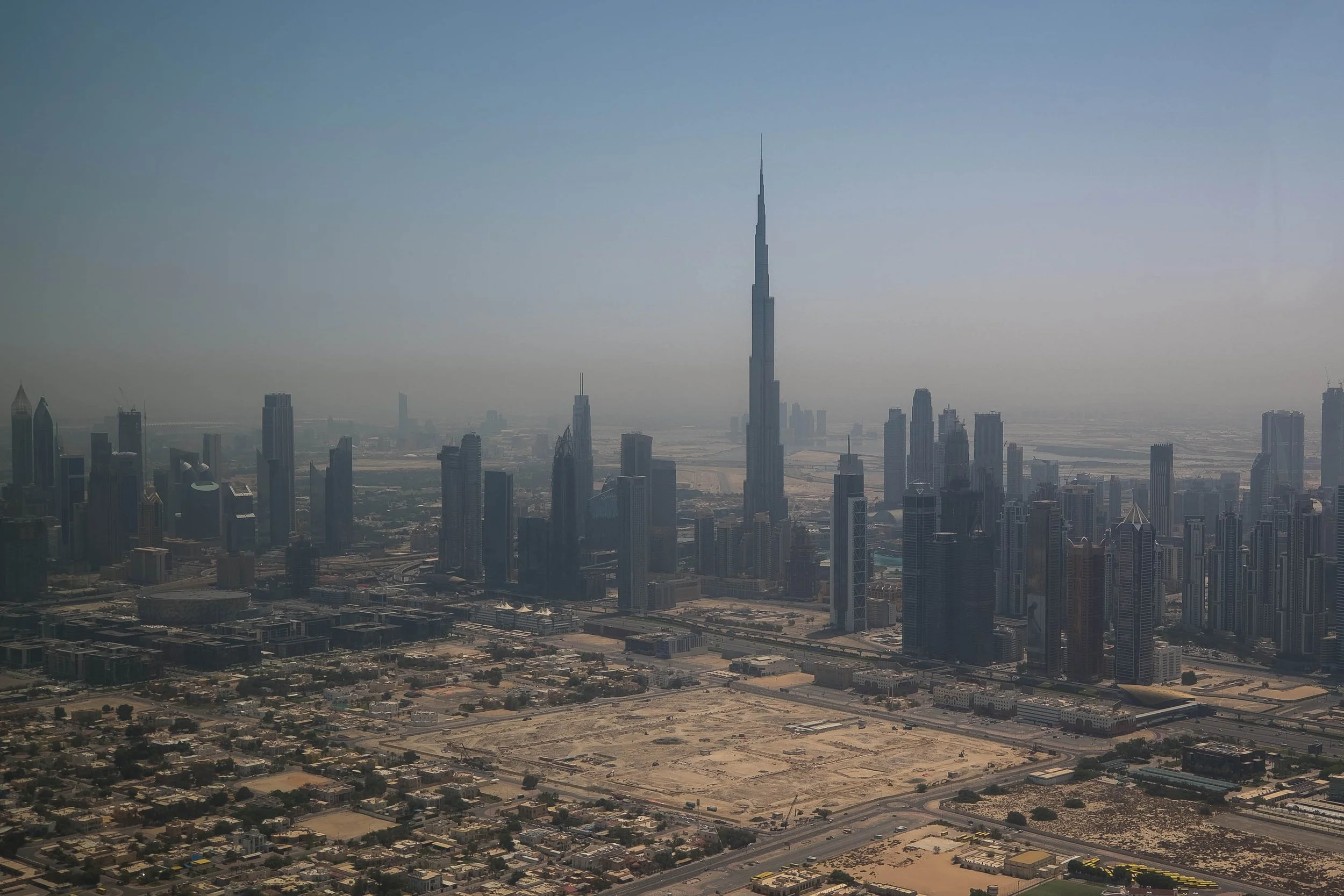 Aerial view of Dubai skyline with Burj Khalifa tower in the center, surrounded by modern skyscrapers and sandy areas in the foreground.