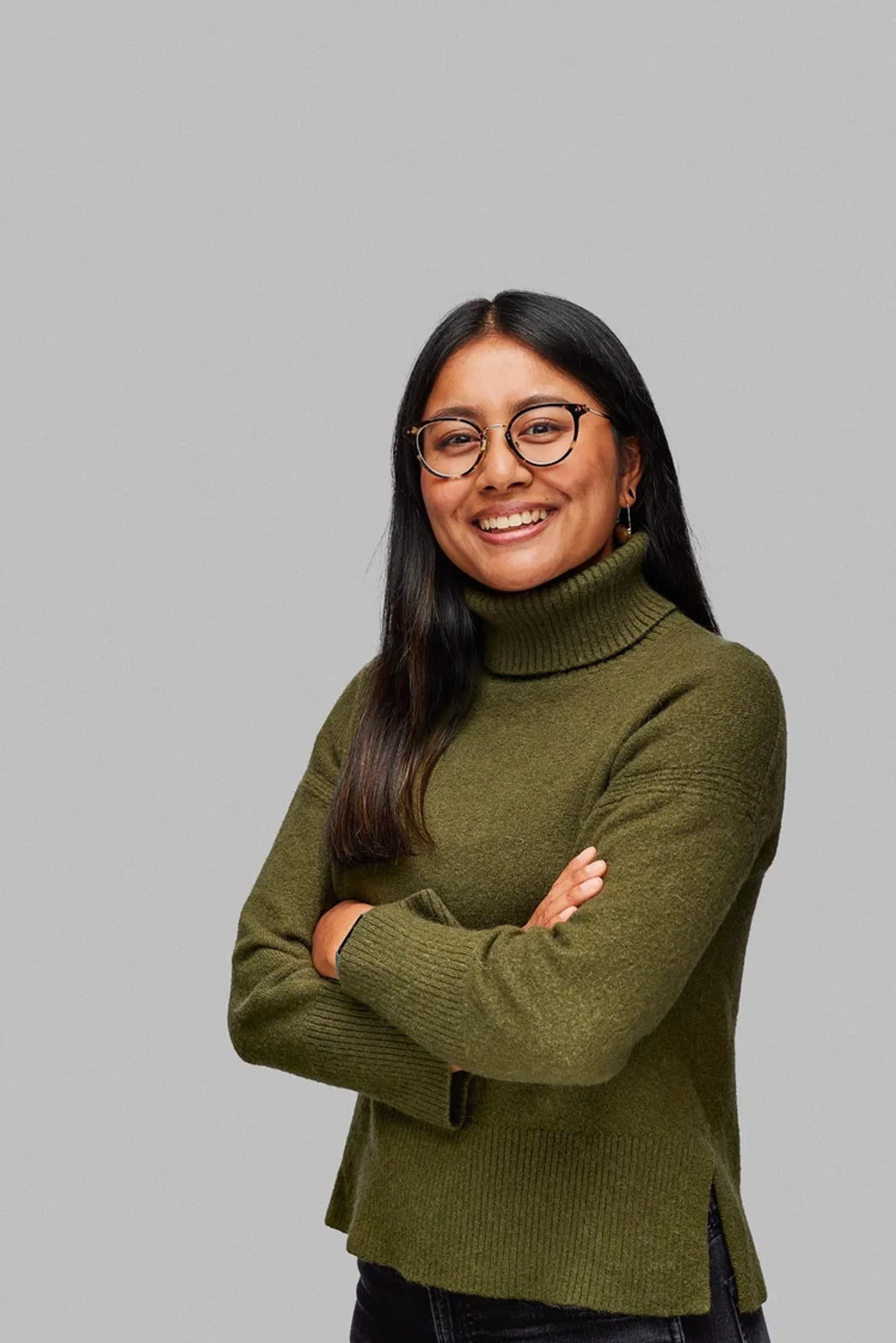 A woman with long dark hair and glasses smiling, wearing a green turtleneck sweater and black pants, standing with her arms crossed against a plain light gray wall.