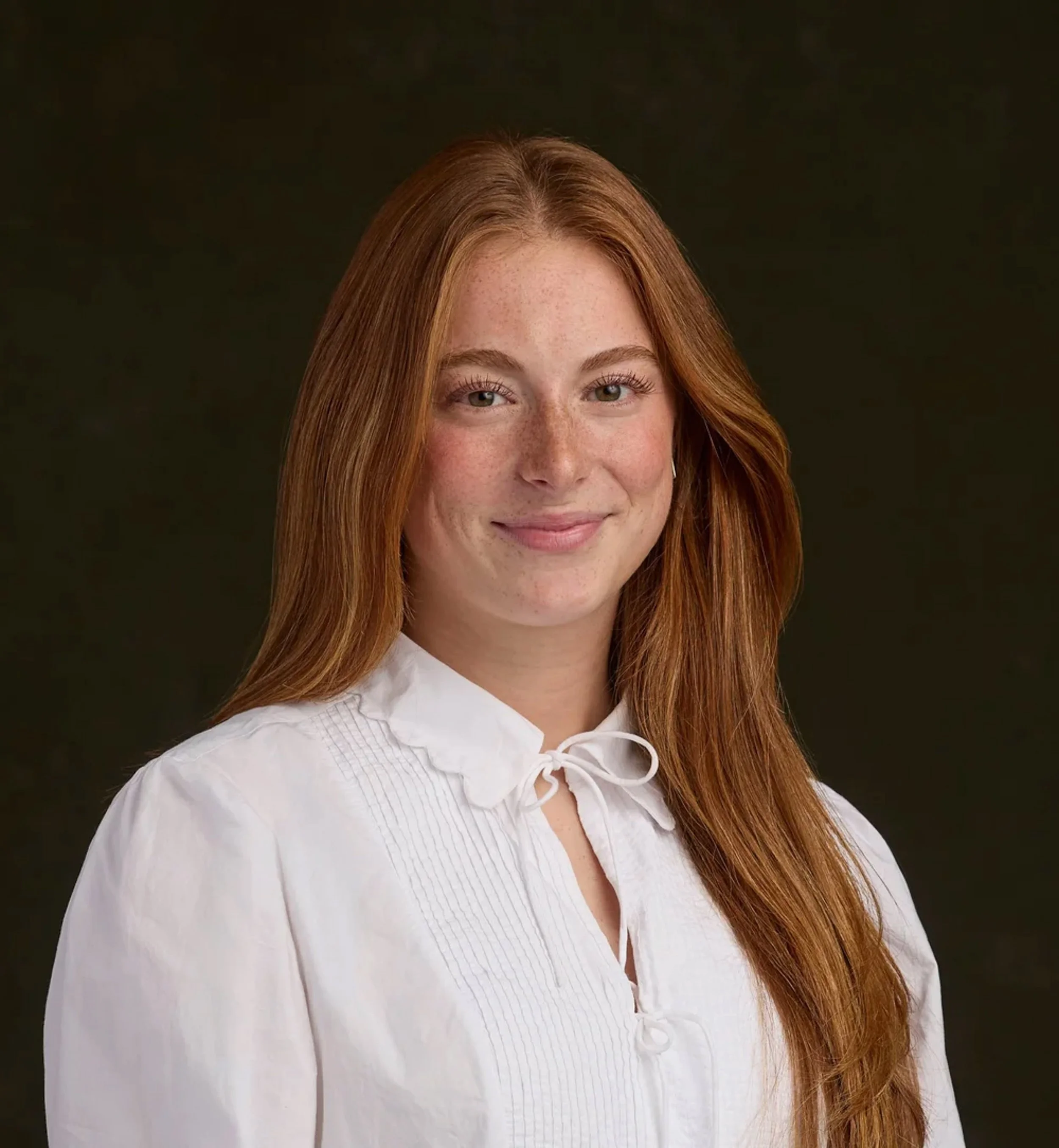 Close-up portrait of a young woman with long, wavy red hair, blue eyes, and light freckles, wearing a white collared shirt, against a dark backdrop.