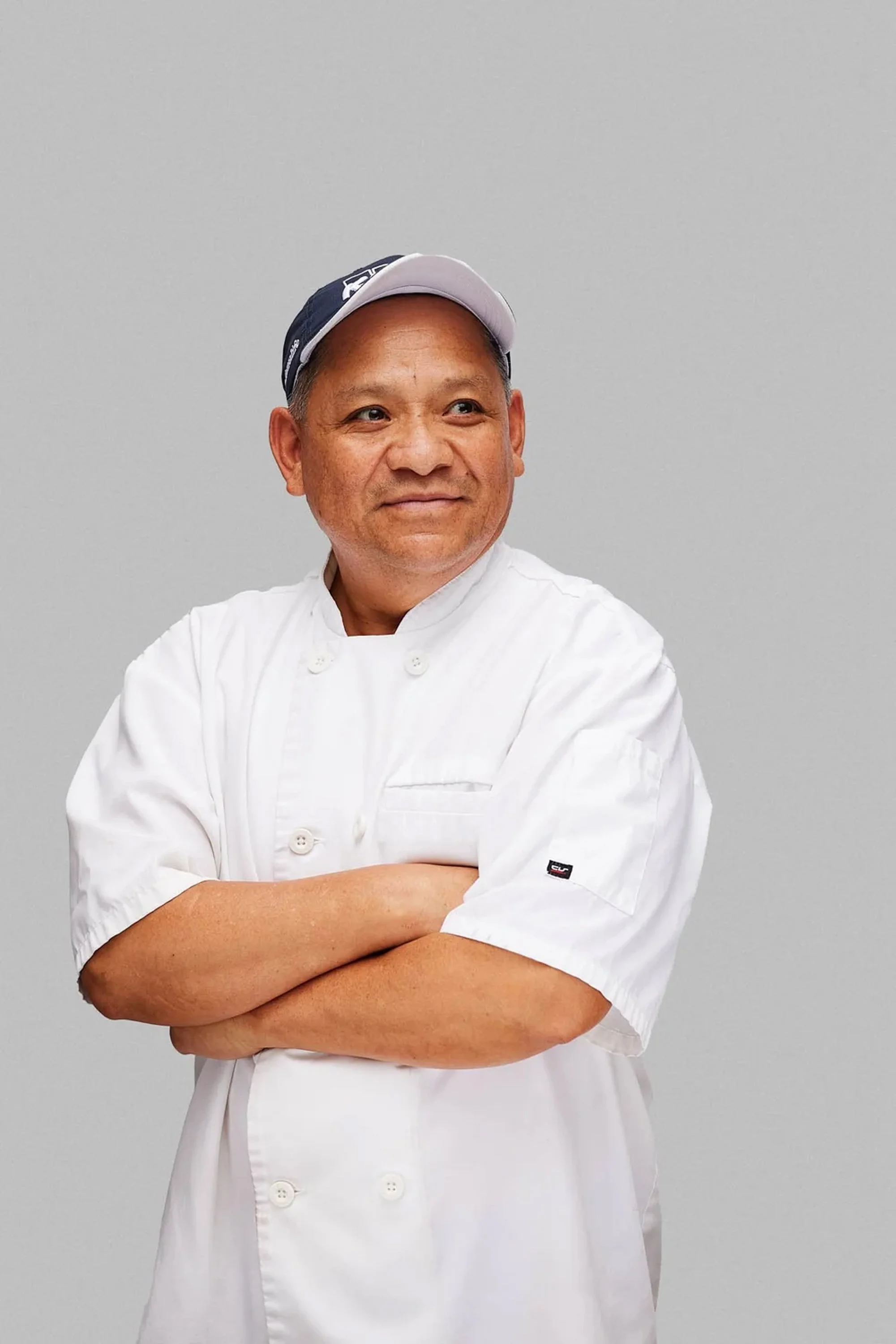 A man wearing a white chef's coat and a black and white cap, standing with his arms crossed against a plain gray background.