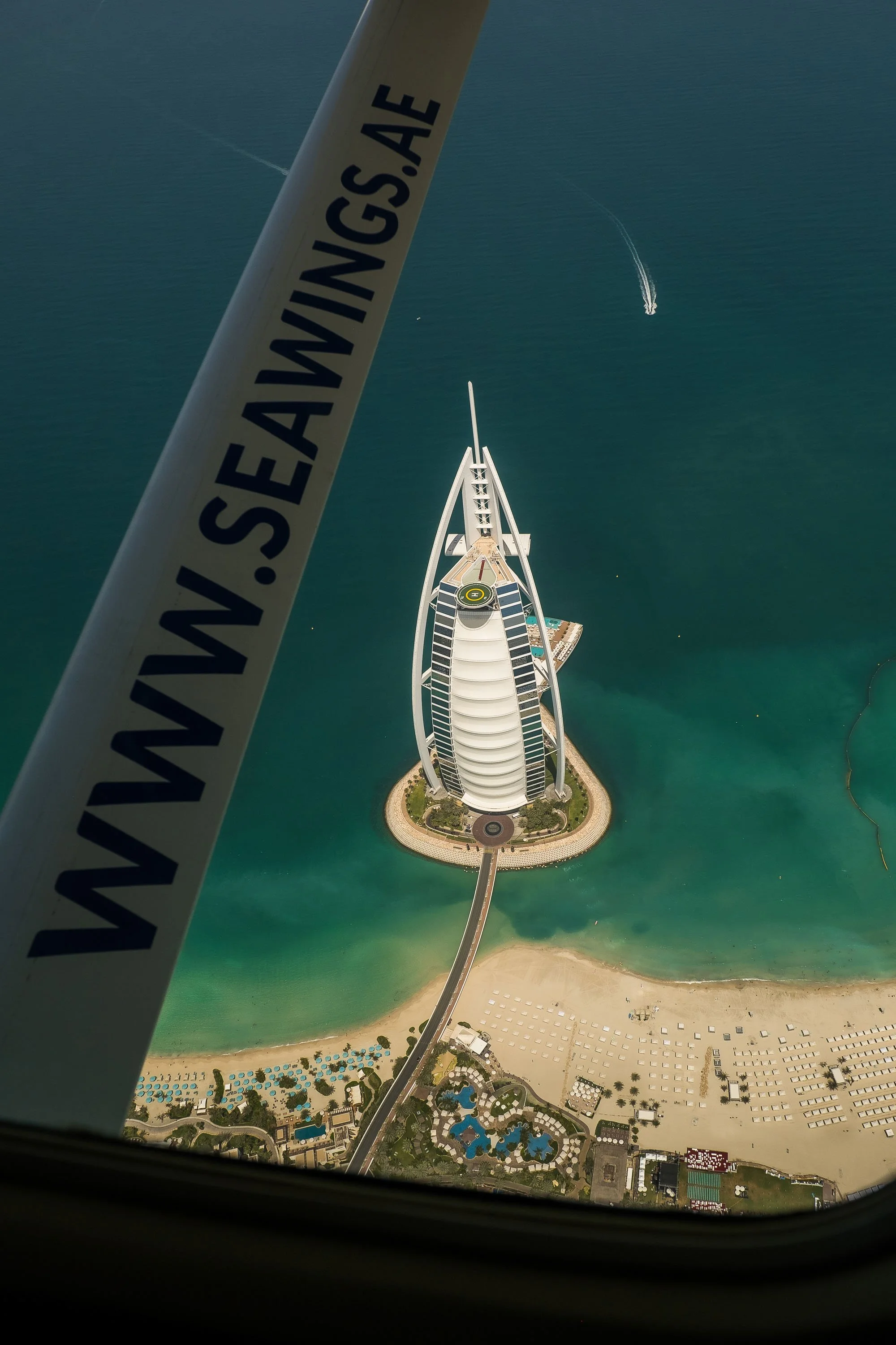 Bird's eye view of the Burj Al Arab hotel in Dubai, seen from a helicopter, with water and beach below and a boat leaving a contrail in the sky.