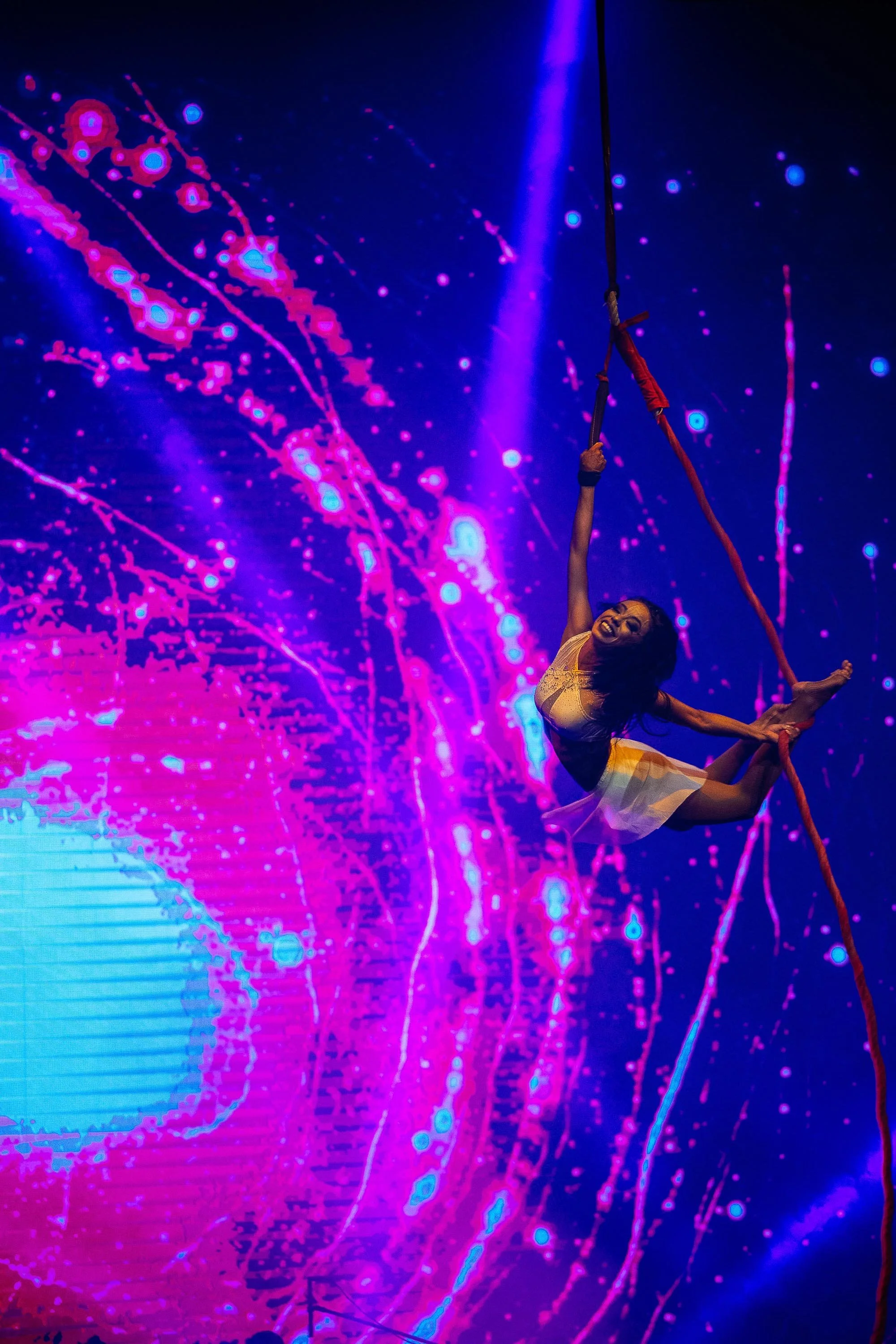 A female acrobat performs on a red rope in front of a vibrant, colorful digital backdrop with pink and purple abstract patterns.