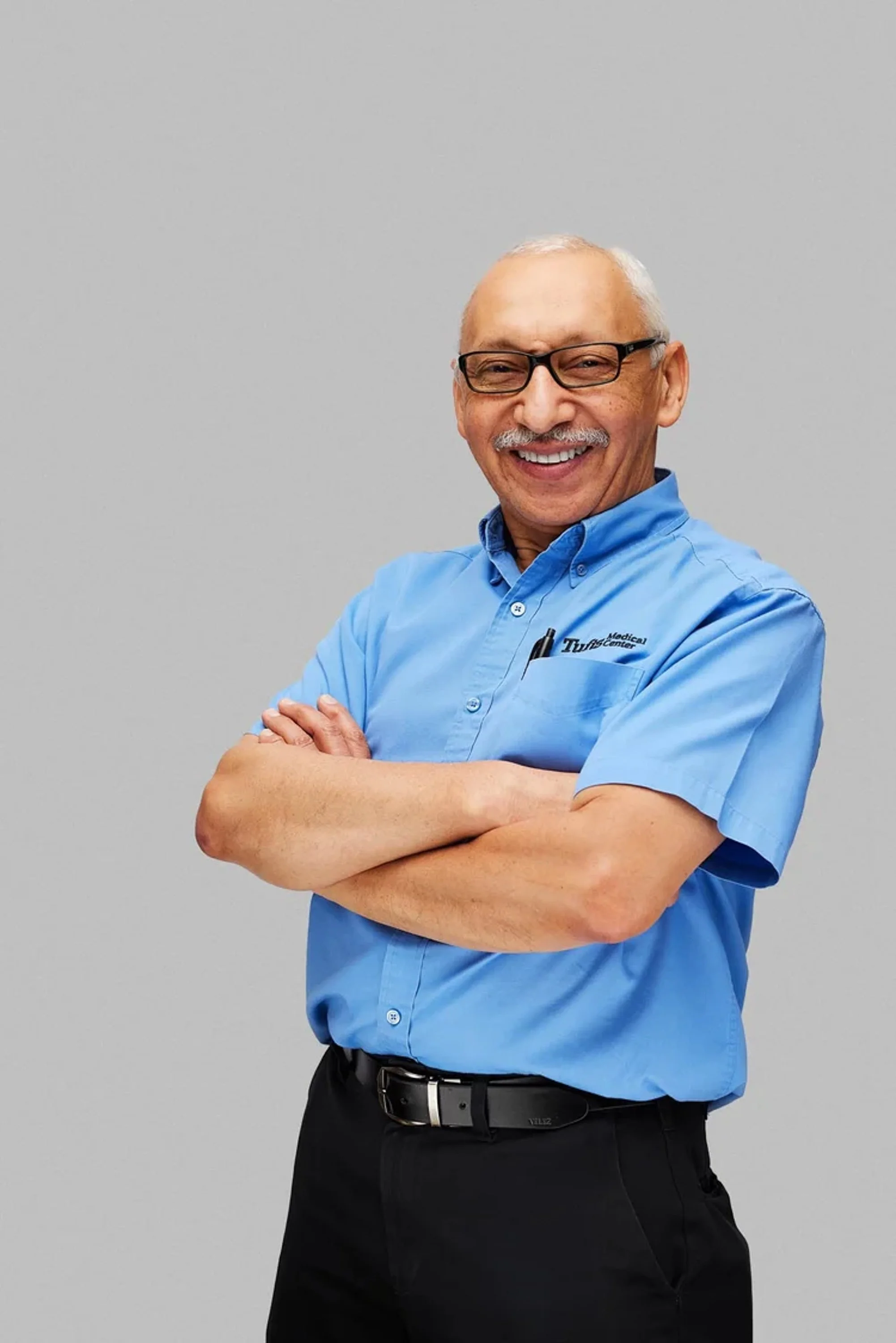 A smiling older man with glasses, bald head, wearing a blue work shirt with a company logo, standing with arms crossed, against a plain gray background.