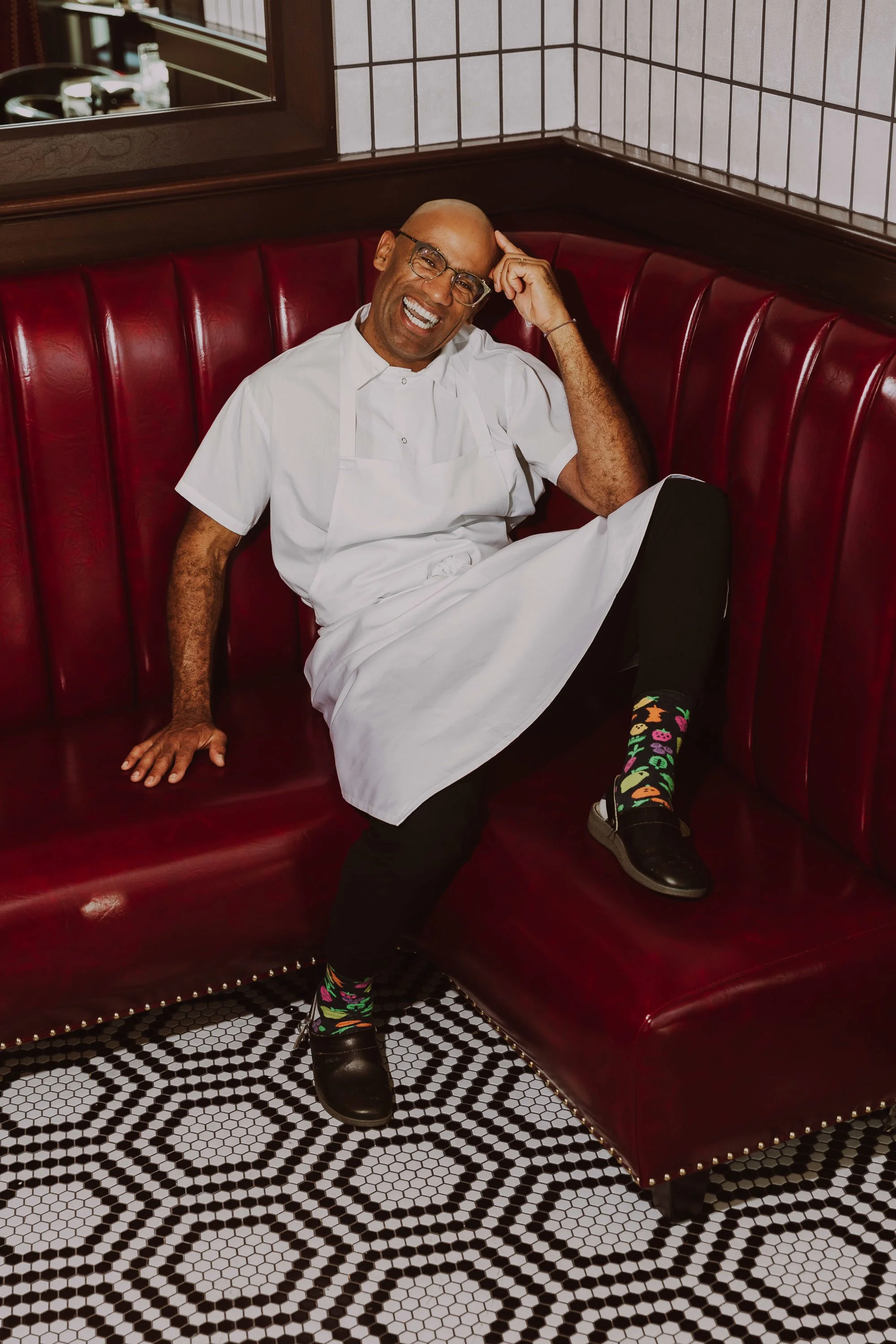 Man sitting on a red leather booth, wearing a white chef's coat, colorful socks with pizza print, and black shoes, smiling and leaning back.
