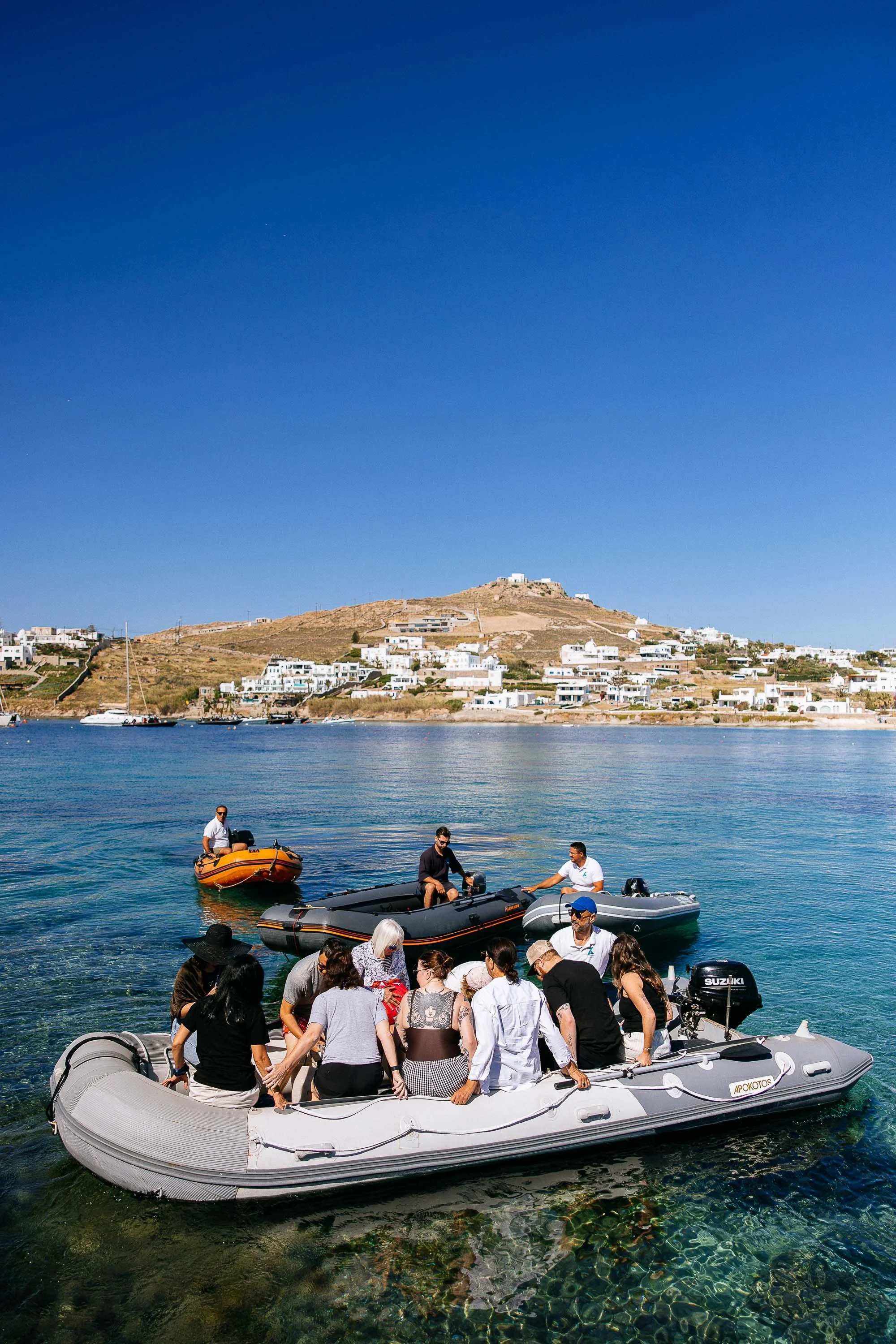 Group of people on a inflatable boat and smaller boats in water, with a hillside town with white buildings in the background on a sunny day.