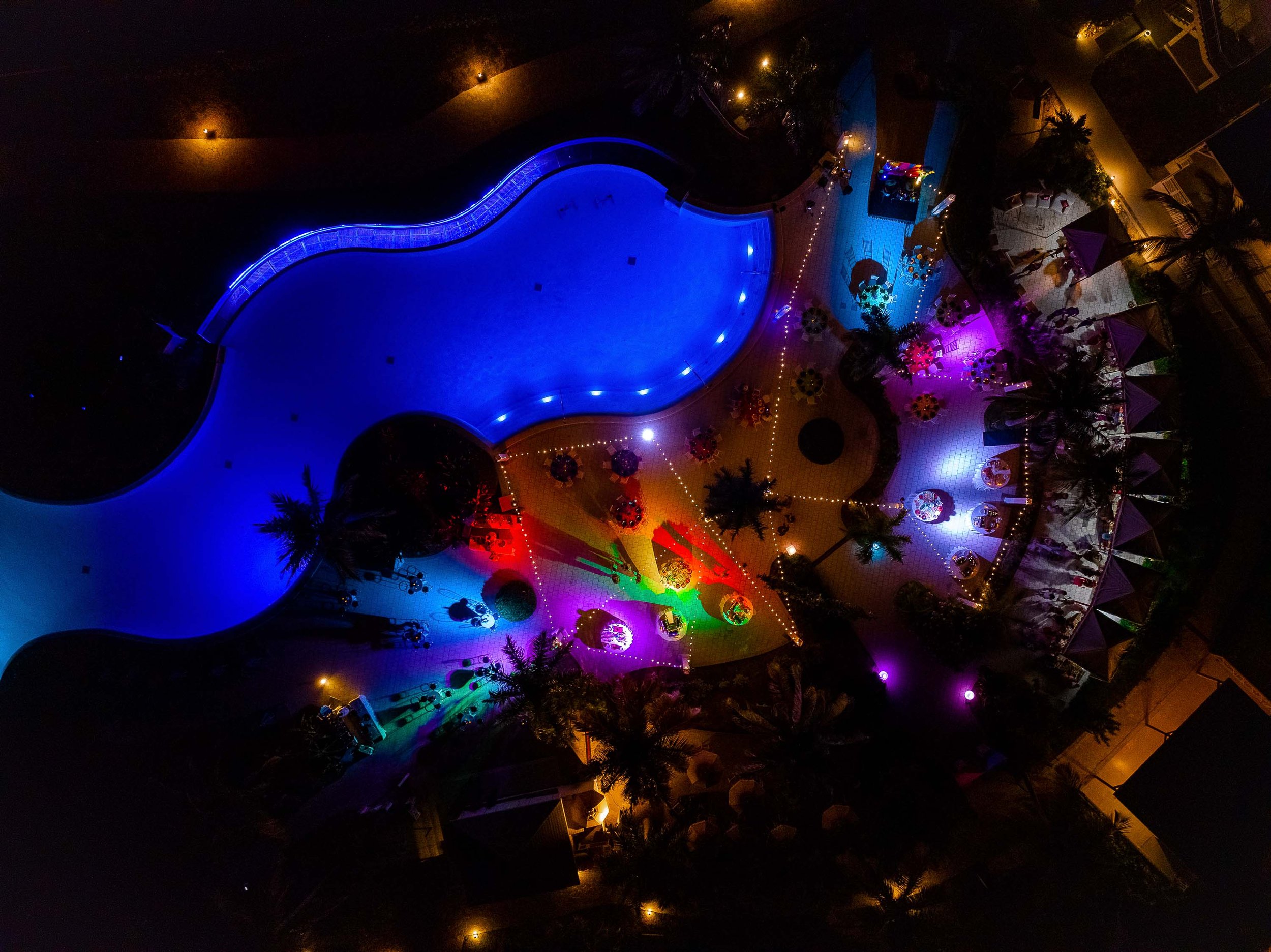 Aerial night view of a swimming pool illuminated with colorful lights, surrounded by lounge chairs and palm trees.