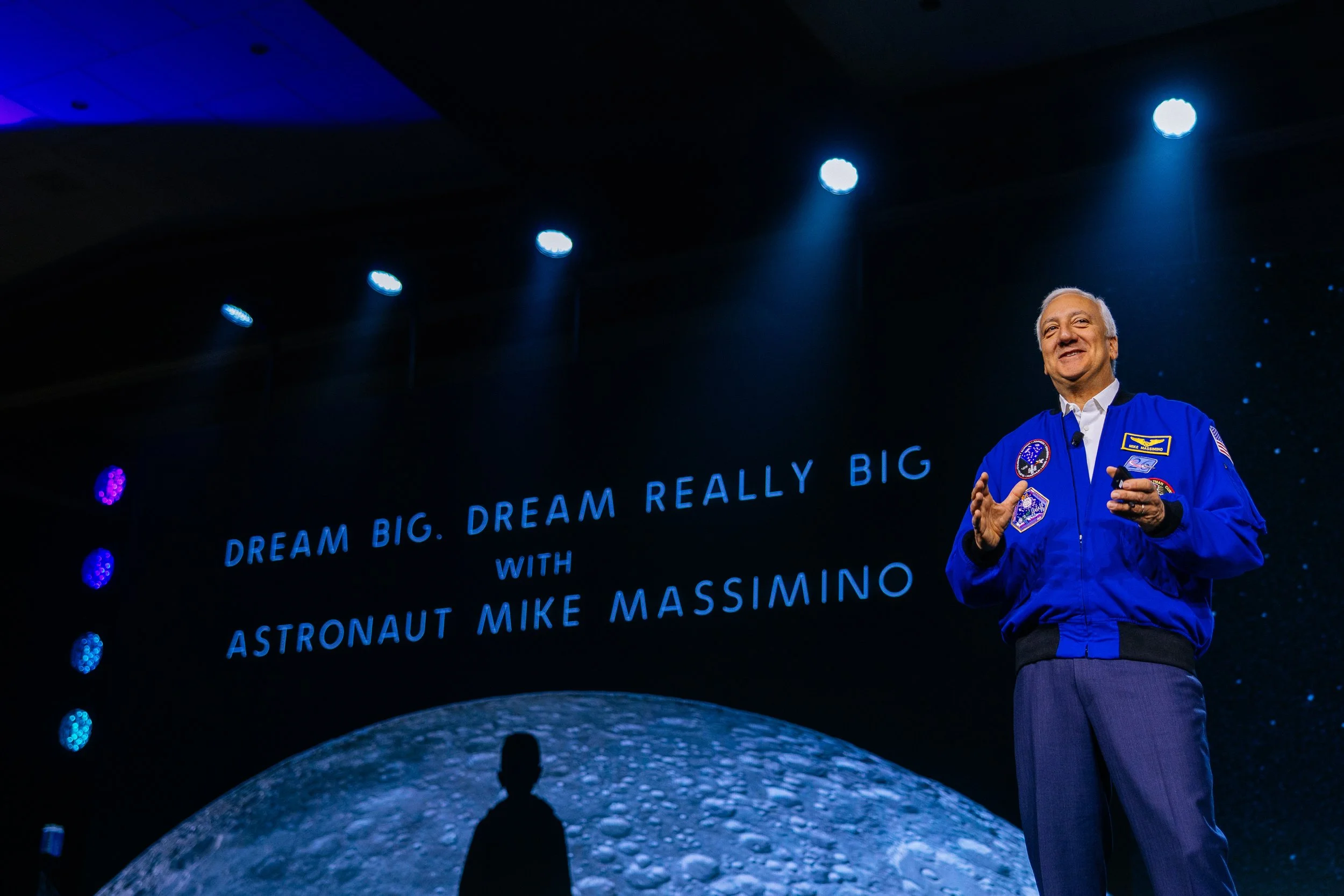 Astronaut Mike Massimino giving a presentation on stage in front of a large screen with the text 'Dream Big. Dream Really Big with Astronaut Mike Massimino.' The background shows a lunar surface and starry sky.