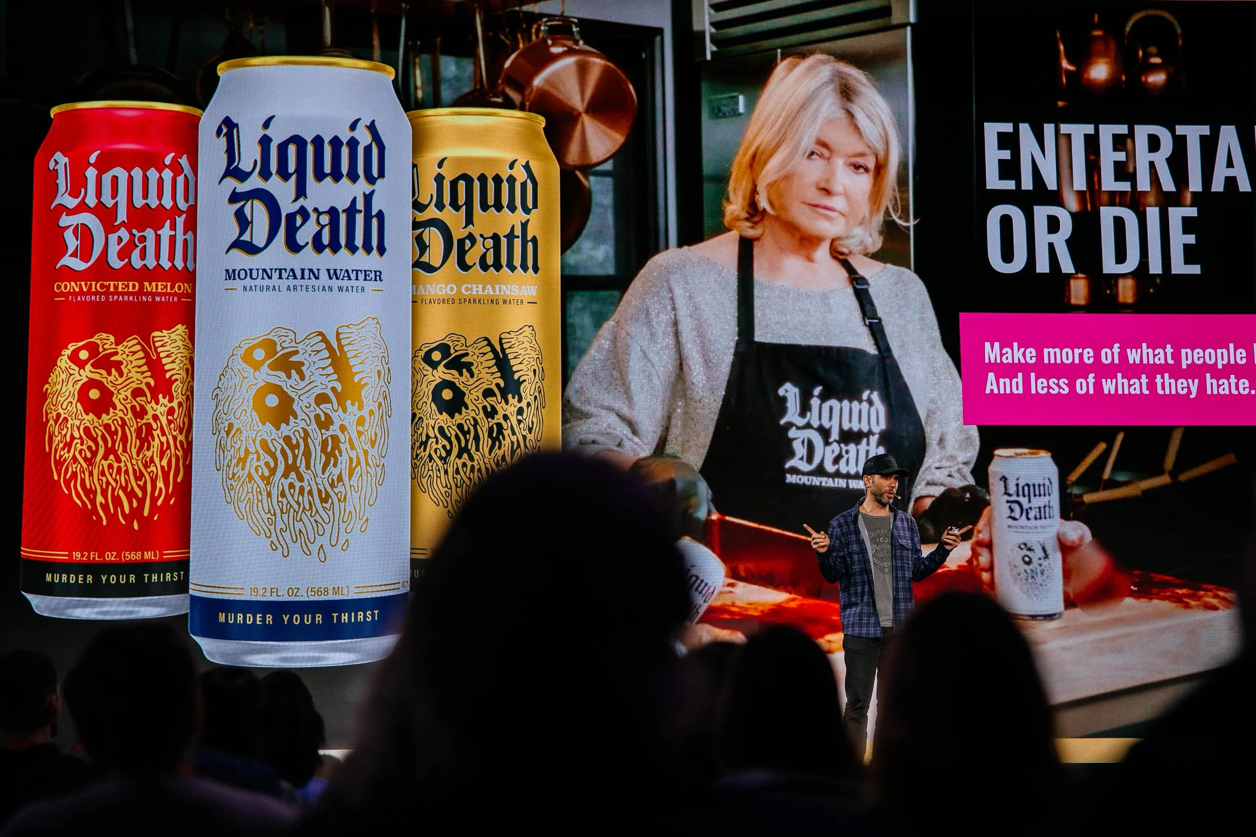 A man is giving a presentation or speech in front of a large screen displaying promotional images of Liquid Death canned water, with the slogan 'Murder Your Thirst' and slogans 'Entertain or Die' and 'Make more of what people want and less of what th