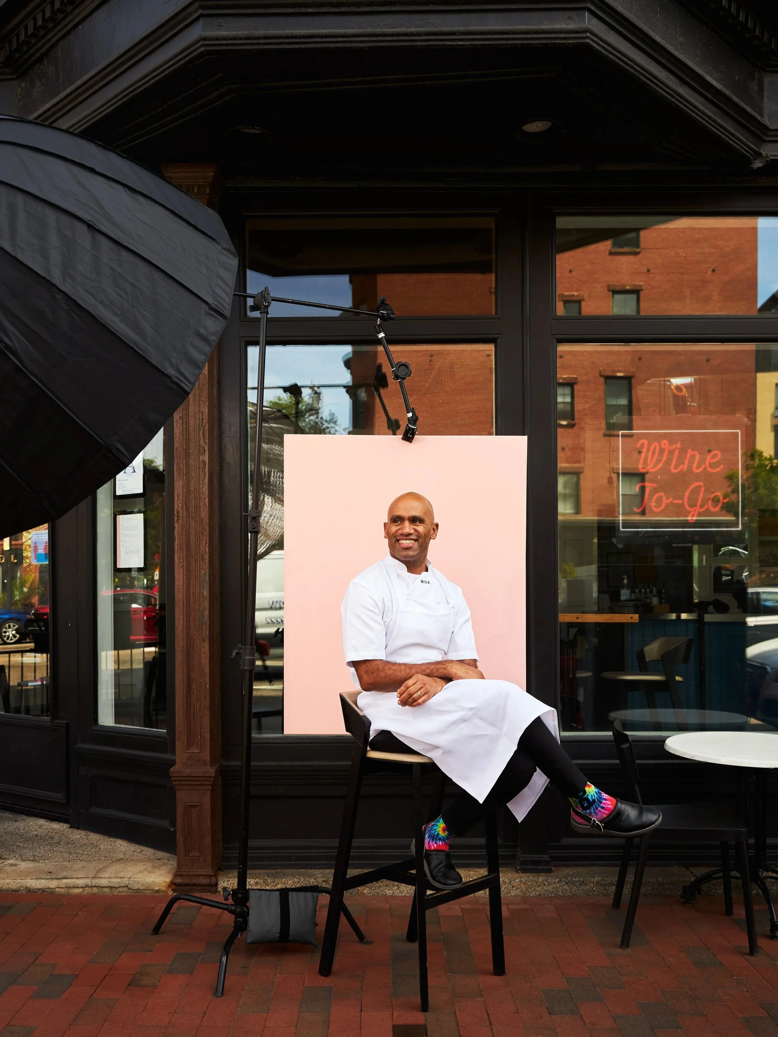 A man in a white chef's coat and black pants with colorful socks is sitting on a chair outside a restaurant. There is a photo backdrop behind him, and a large studio light on the left. The restaurant window has a neon sign that says "Wine To-Go."