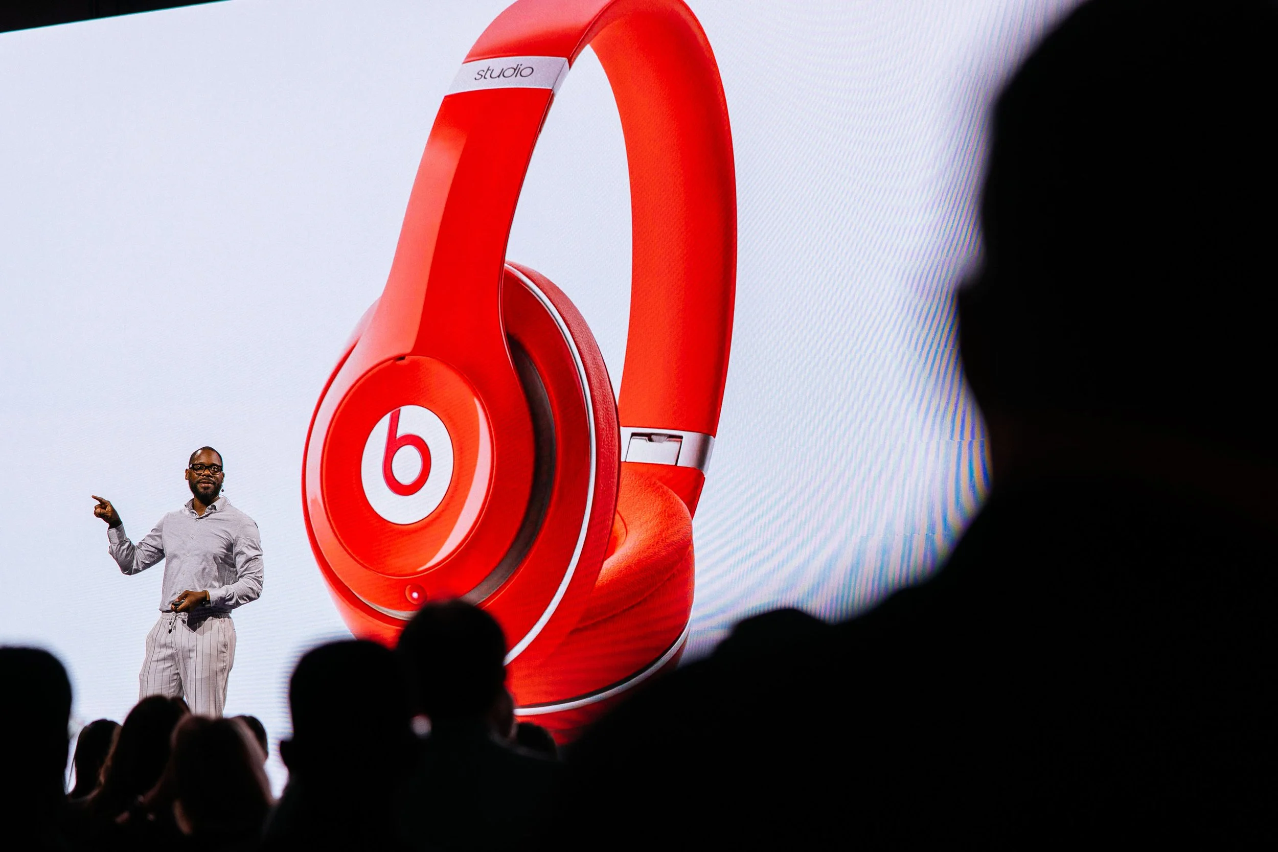 A man giving a presentation on stage with a large screen behind him showing a red pair of Beats headphones with a white 'b' logo.