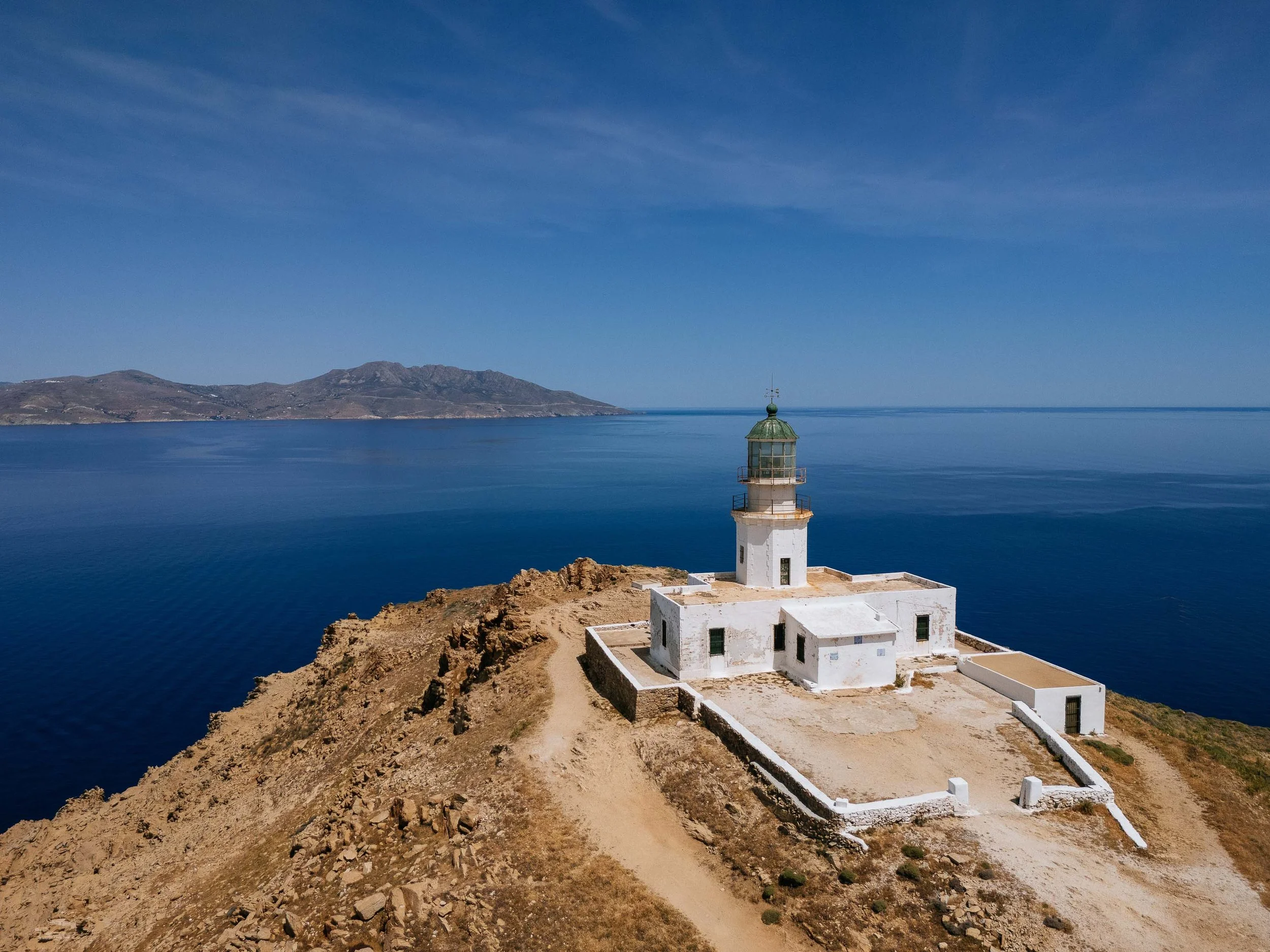 A white lighthouse on a small island with rocky terrain, overlooking a calm, blue sea with a distant mountainous island under a clear blue sky.