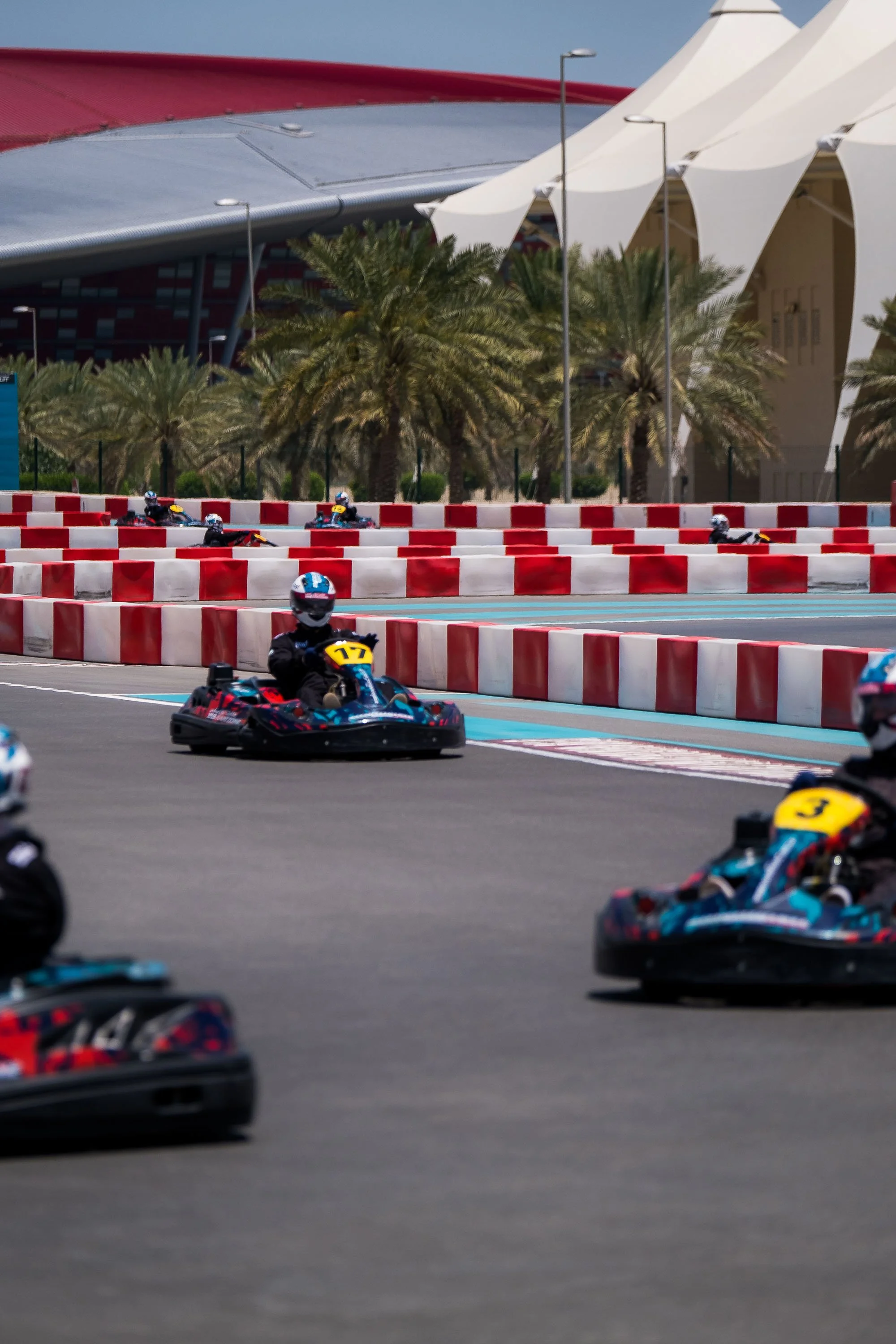 Multiple people in go-karts racing on a track with red and white barriers, palm trees, and modern architecture in the background.