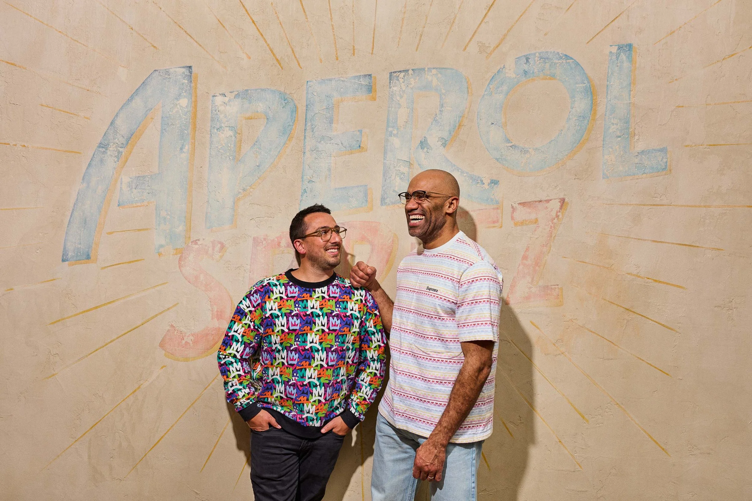 Two men standing in front of a painted wall that says 'ABREO' with rays emanating from the lettering. One man is wearing a colorful patterned sweatshirt, glasses, and has his hands in his pockets. The other man is wearing a striped t-shirt, glasses, 