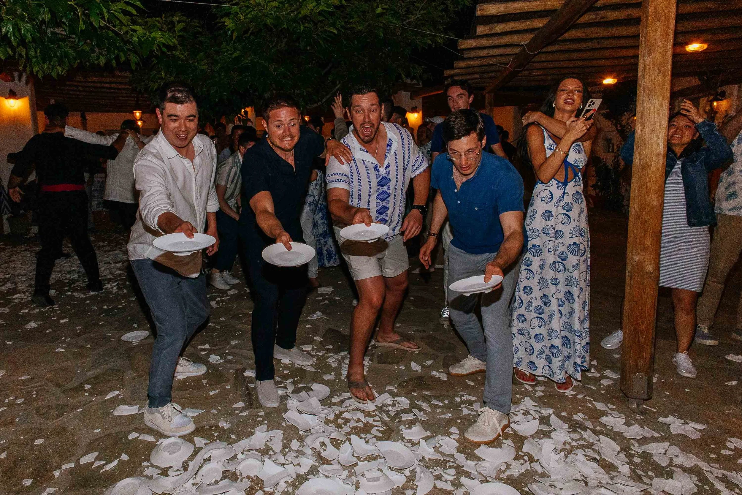 Group of people dancing and celebrating outdoors at night, with broken plates on the ground, some holding plates, some taking photos, under a wooden canopy.