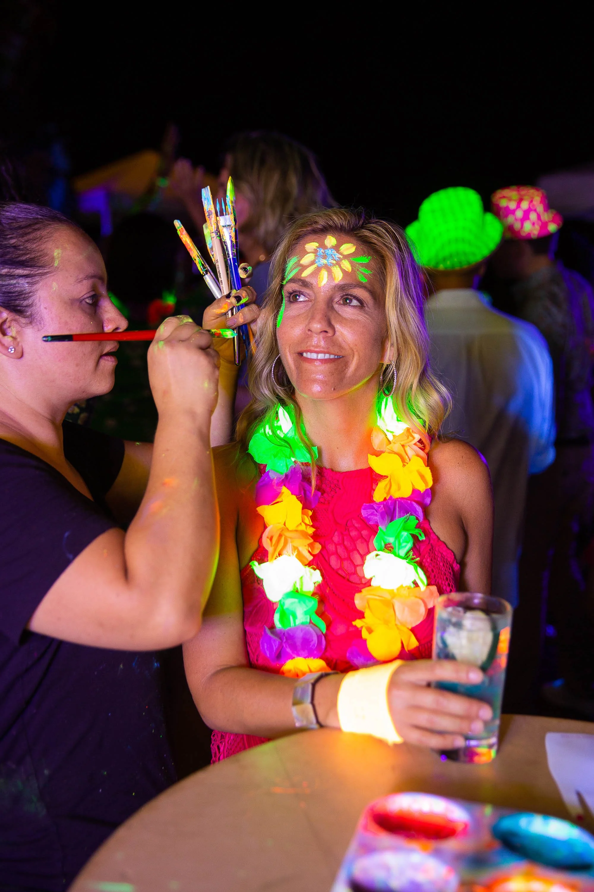 Woman at a party receiving face paint on her forehead, wearing a colorful flower lei, holding a drink, with others in fun hats and glow-in-the-dark accessories in the background.