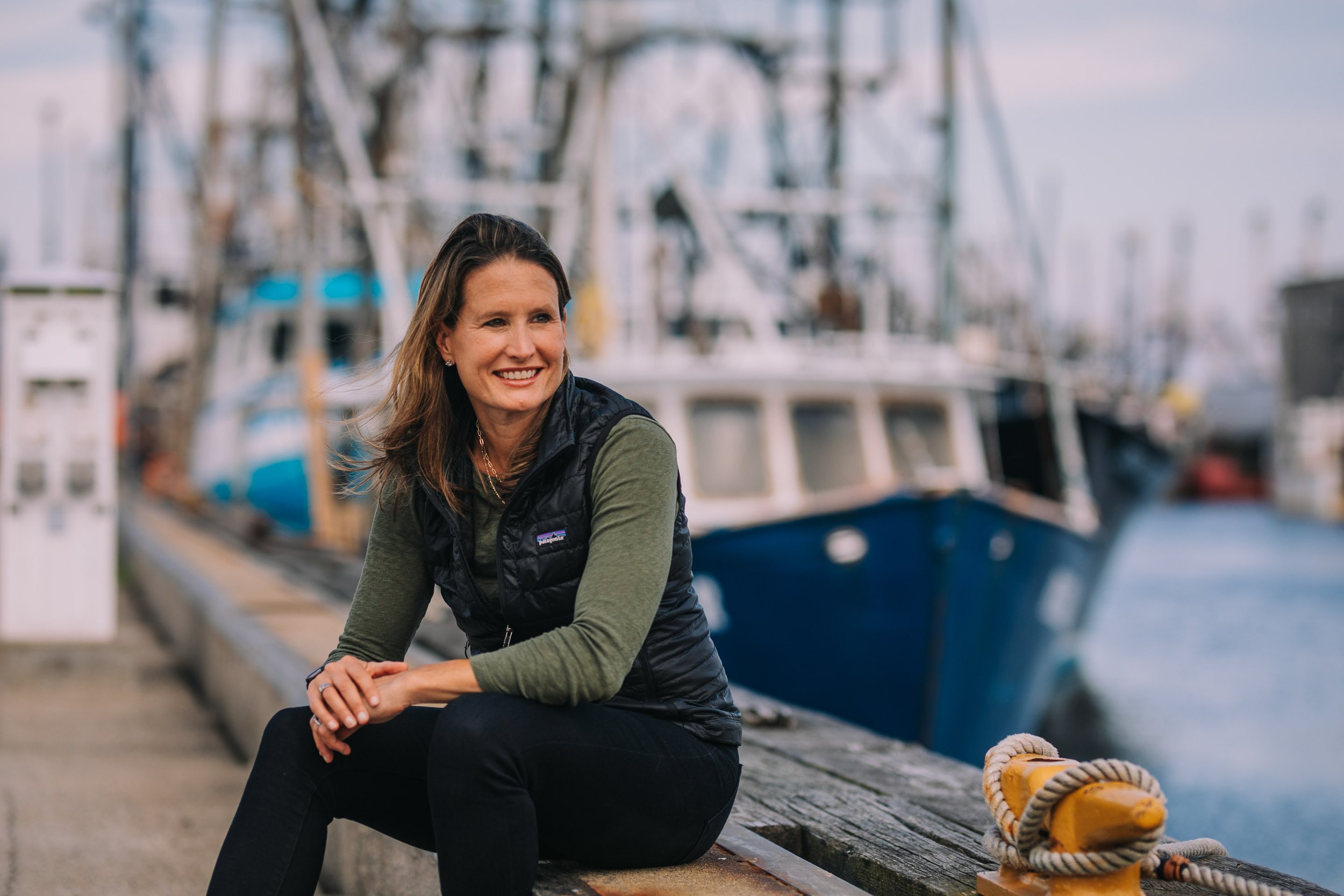 A smiling woman with shoulder-length brown hair sitting on a dock near boats, wearing a dark vest over a long-sleeve shirt, with a marina in the background.