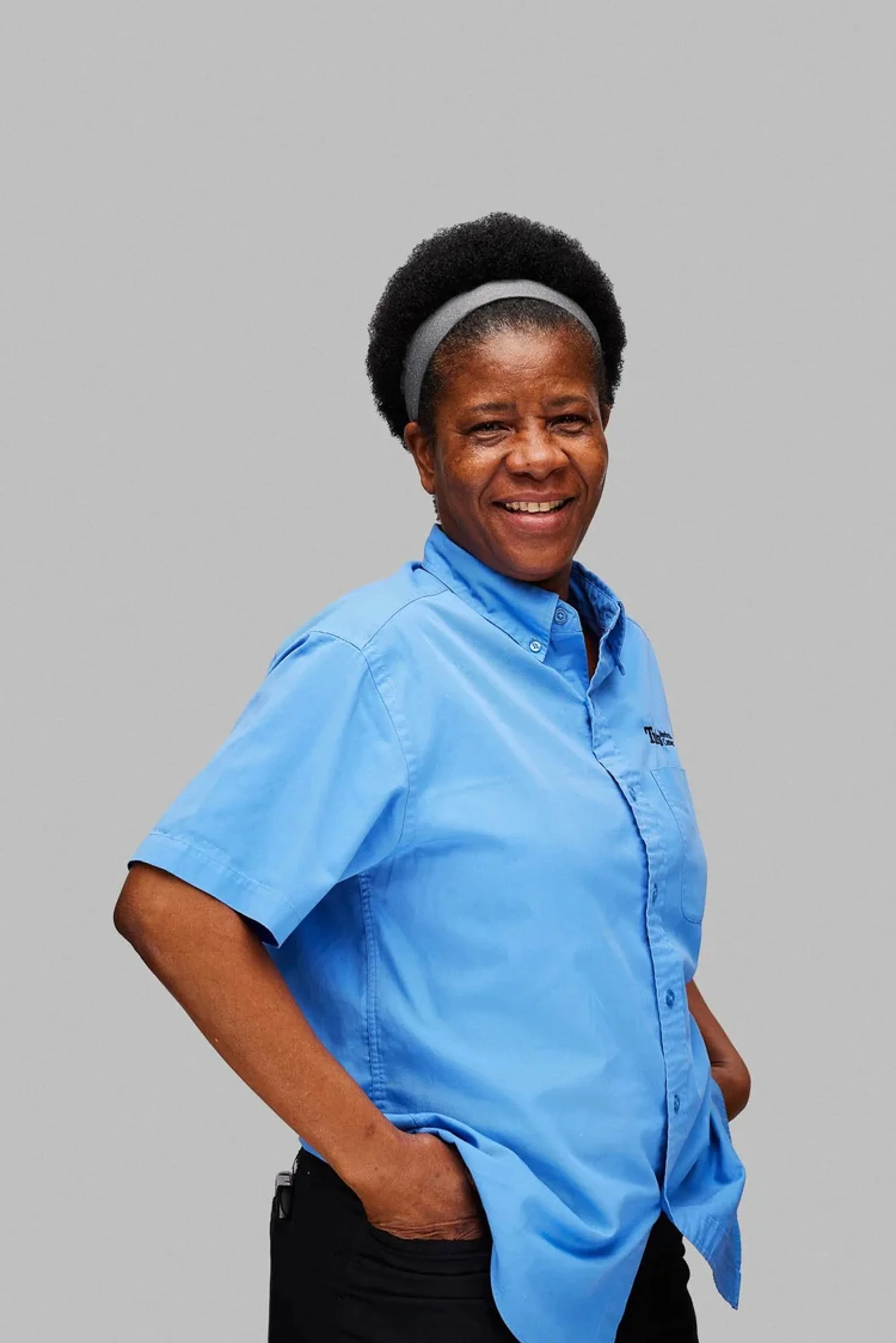 A woman with dark skin, short curly hair, and a gray headband, smiling and wearing a blue work shirt and black pants, standing against a plain gray background.