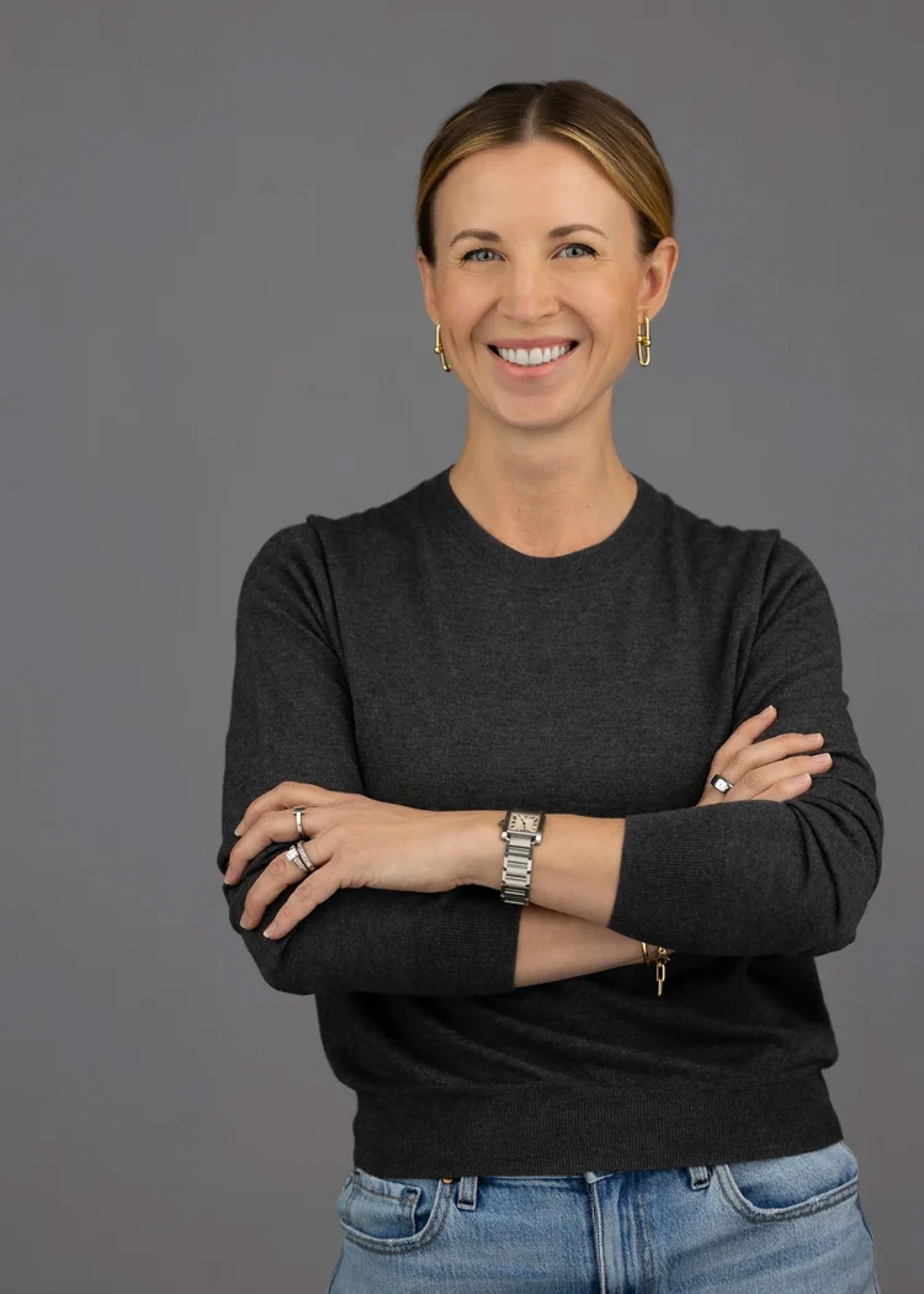A woman with short, light brown hair and blue eyes smiling, wearing a black long-sleeve shirt, gold earrings, a watch, and rings, standing against a gray background with crossed arms.