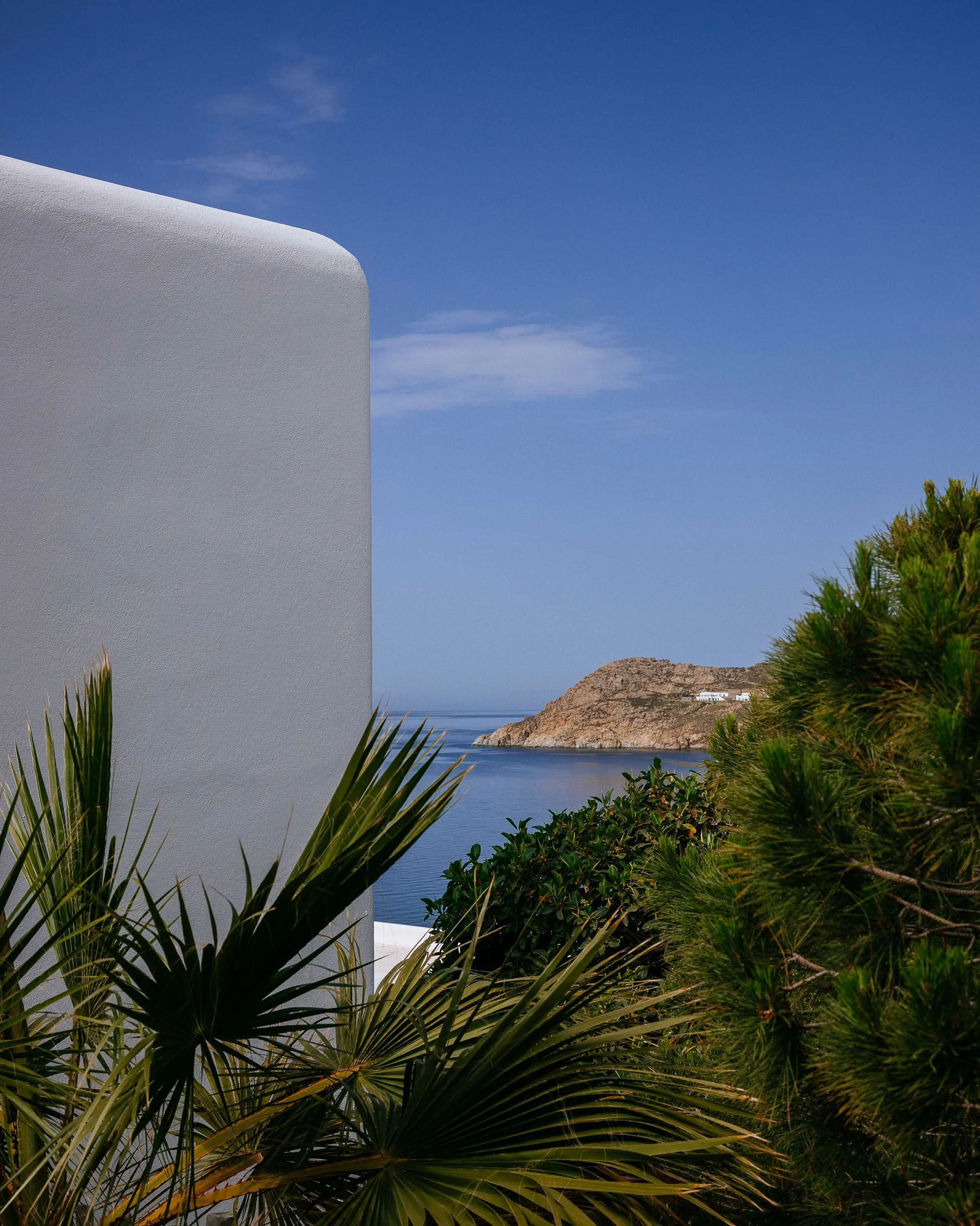 White building corner with lush green palm and pine trees in foreground, blue sea and rocky island in background under a partly cloudy sky.