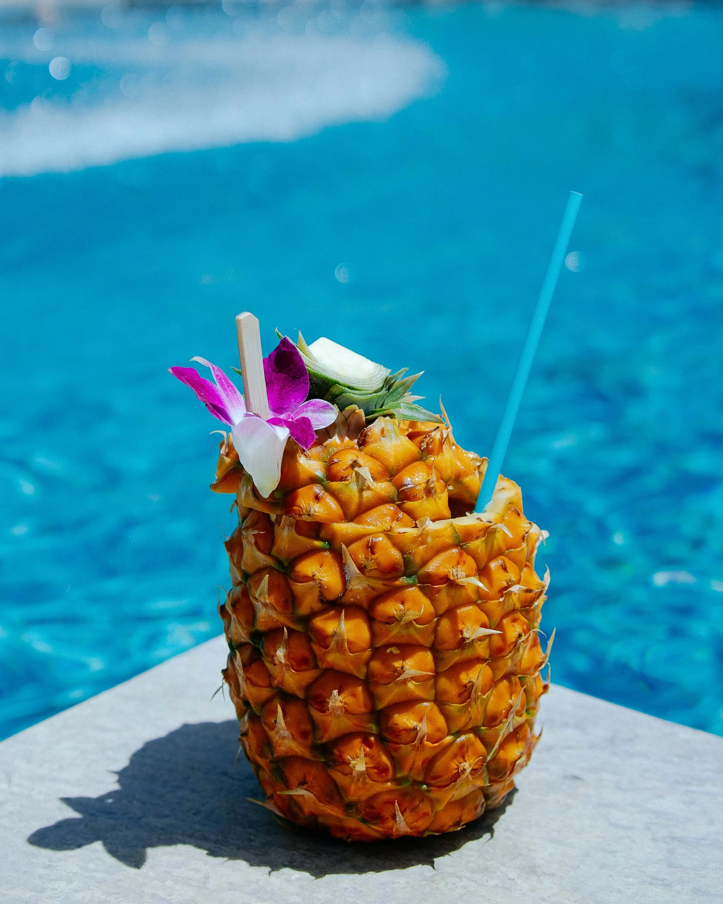 A pineapple drink with a straw, garnished with an orchid flower, a slice of pineapple, and a decorative stick, placed on a concrete surface near a pool.