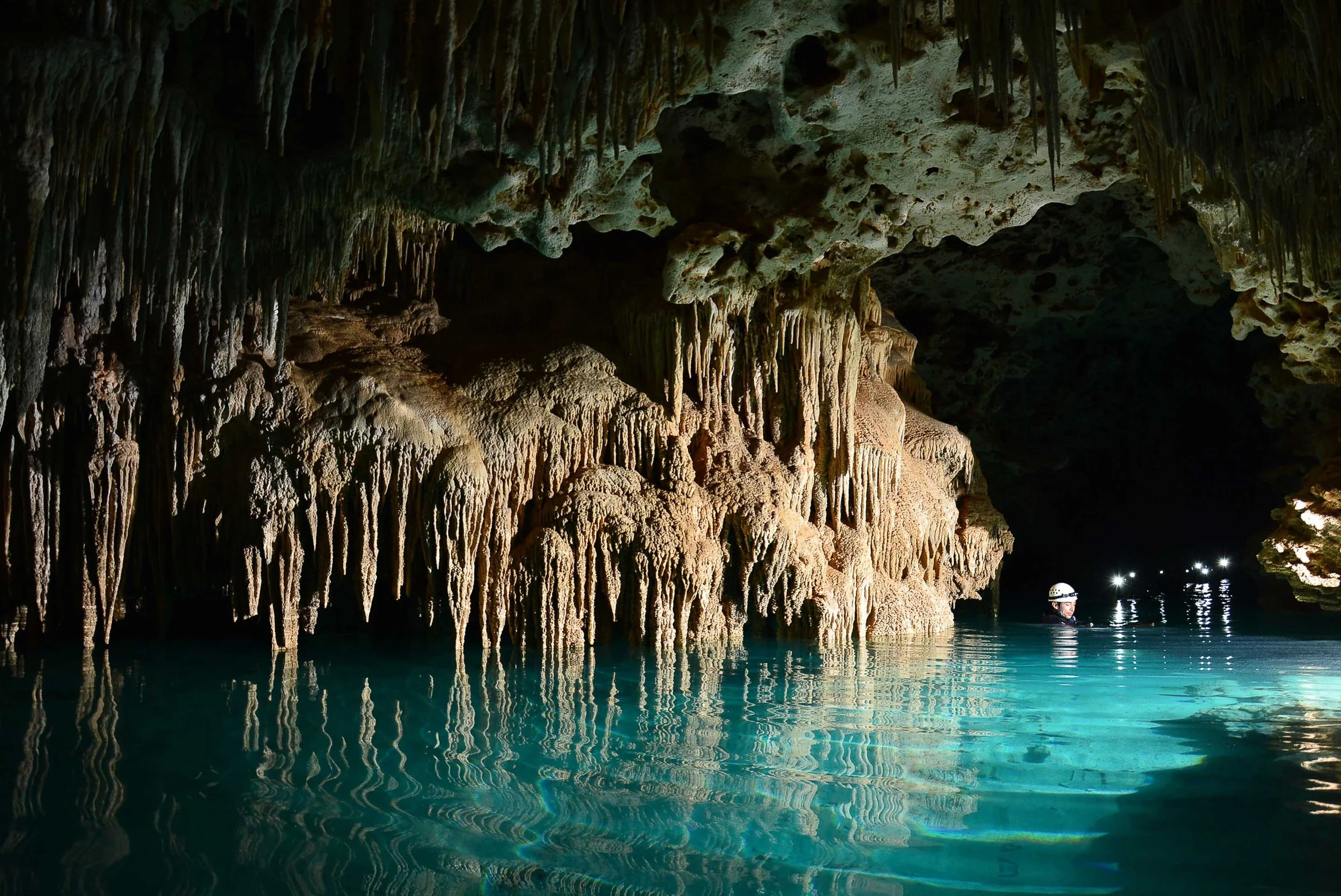 A person wearing a helmet exploring a large underground cave with stalactites hanging from the ceiling and turquoise water reflecting the cave formations.