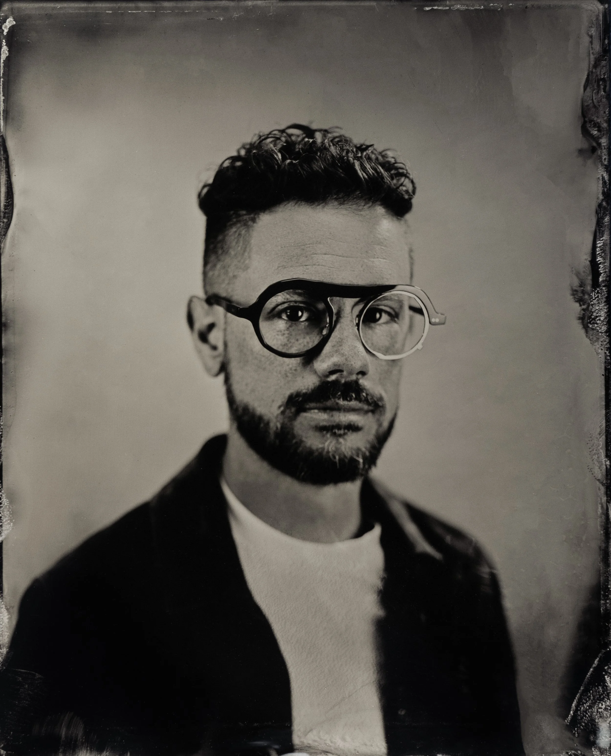 Black and white portrait of a man with short, curly hair, a beard, wearing glasses, a white t-shirt, and a dark jacket.