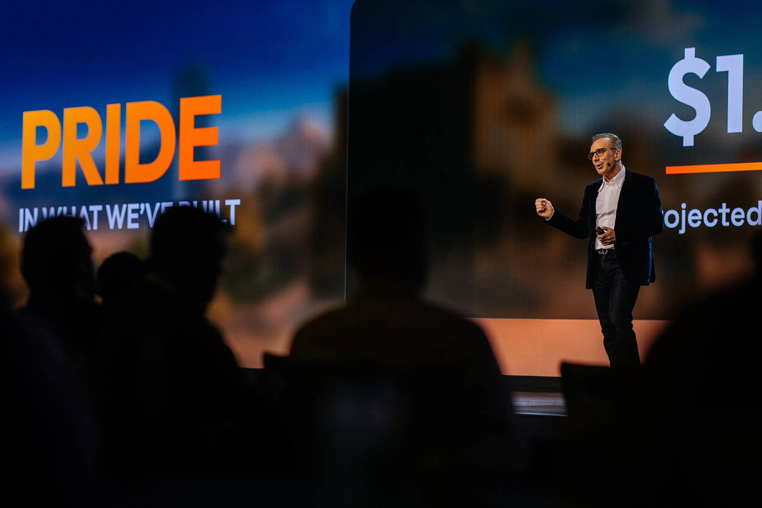 A man giving a presentation on stage with a large screen behind him displaying the words 'PRIDE' and financial figures, while an audience watches in silhouette.