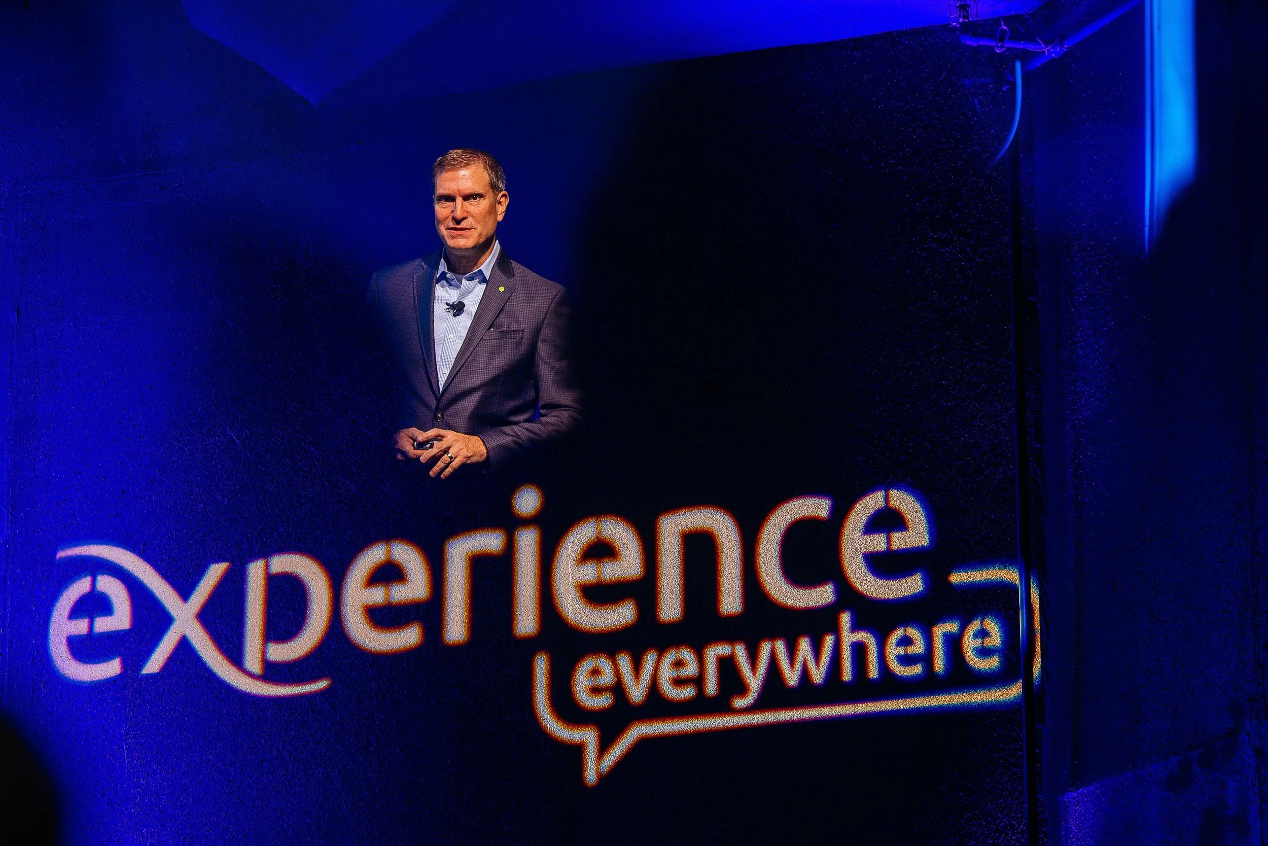 A man in a dark suit standing in front of a black wall with the words 'experience everywhere' projected in neon lights.