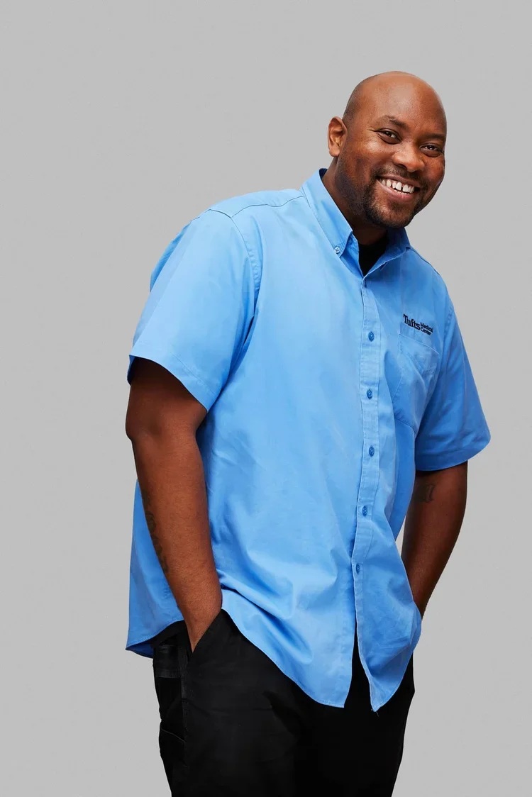 Man in light blue short-sleeve button-up shirt and black pants smiling, standing with hands in pockets against gray background.
