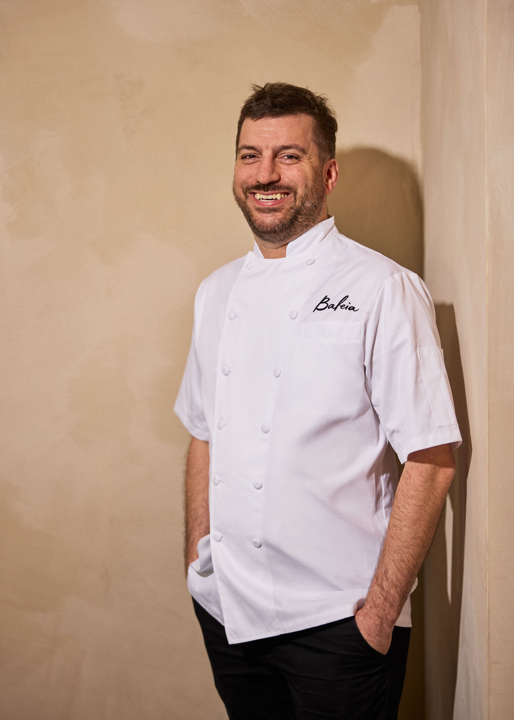 A male chef wearing a white chef's jacket with the name 'Bakeia' embroidered on it. He has short brown hair, a beard, and is smiling while standing against a beige wall with his hands in his pockets.