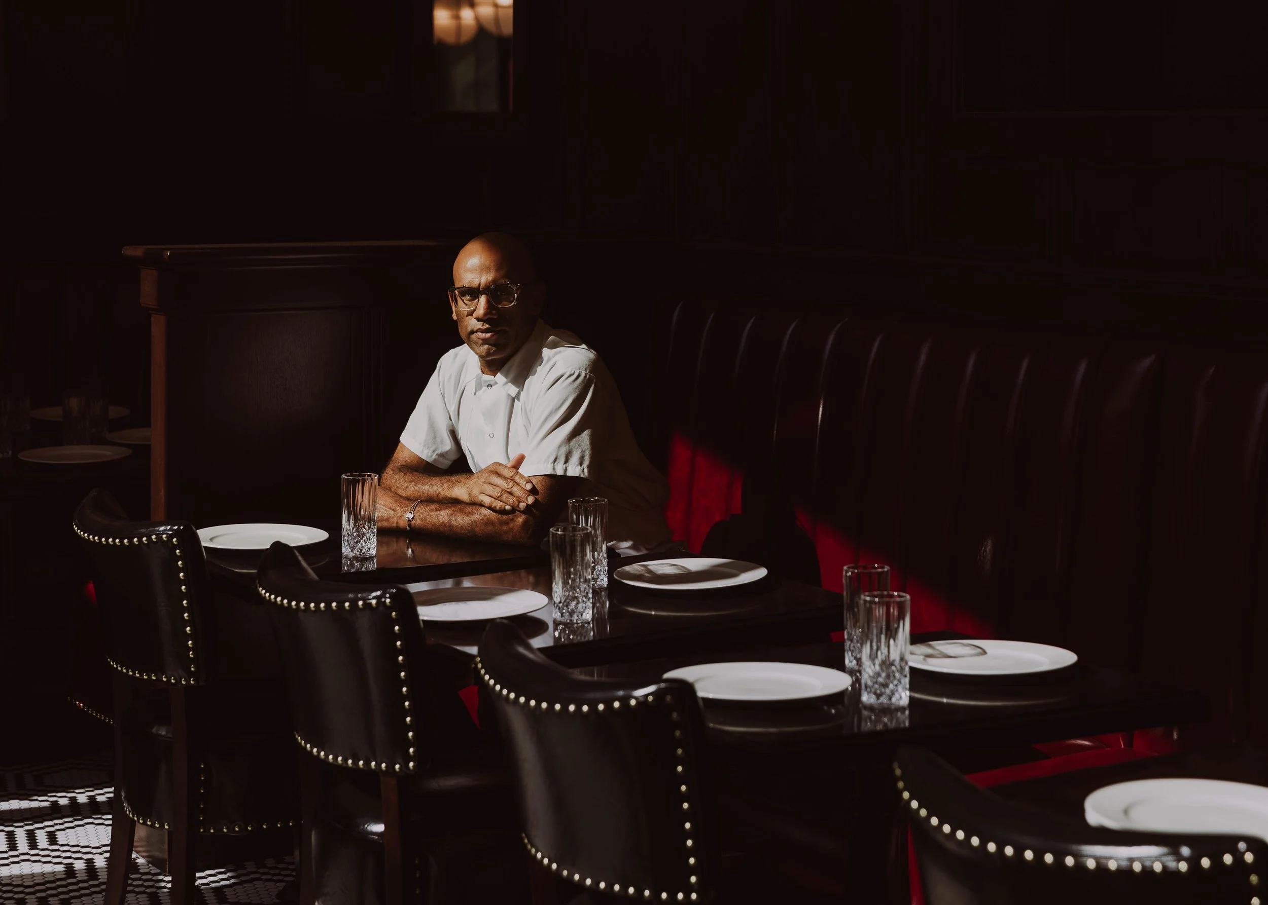 A man with glasses and a white shirt sitting alone at a dining table in a dimly lit restaurant, with empty chairs and glasses on the table.