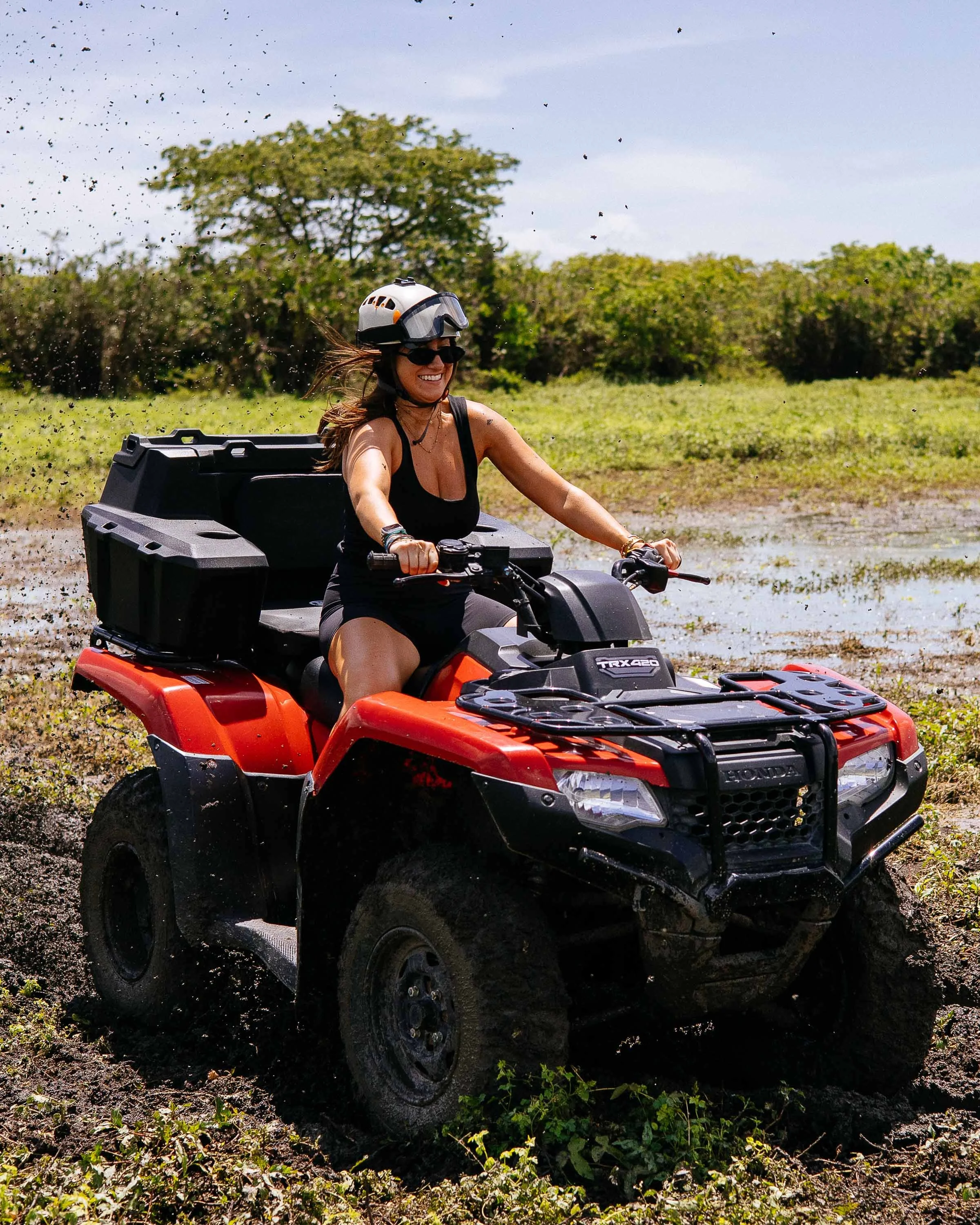 A woman riding a red Honda all-terrain vehicle through a muddy field, wearing a helmet and sunglasses, smiling.