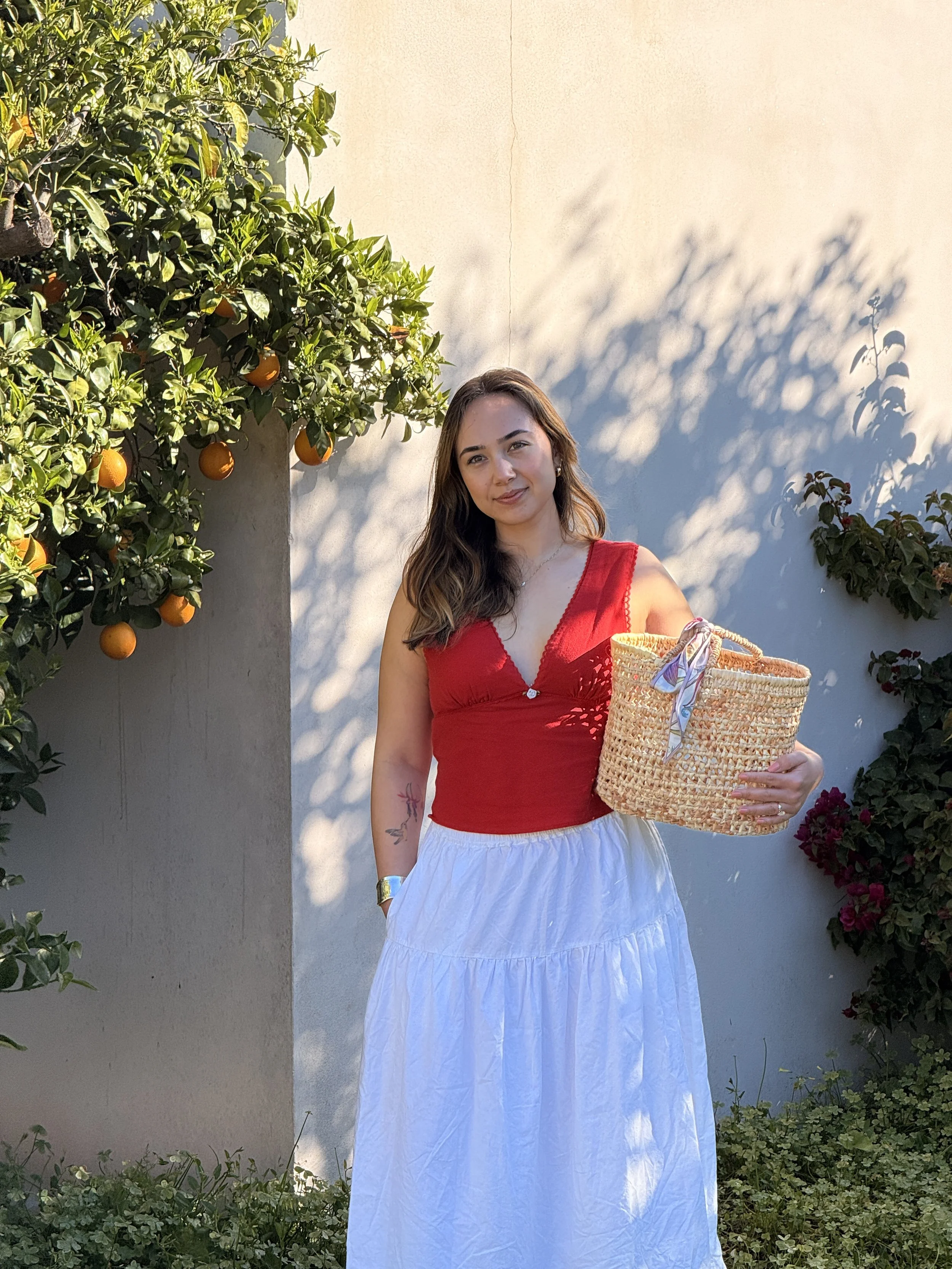 Founder of Solenne. A woman in a red sleeveless top and white skirt standing outdoors next to an orange tree and white wall, holding a woven basket with a colorful scarf. She has wavy brown hair and is smiling.