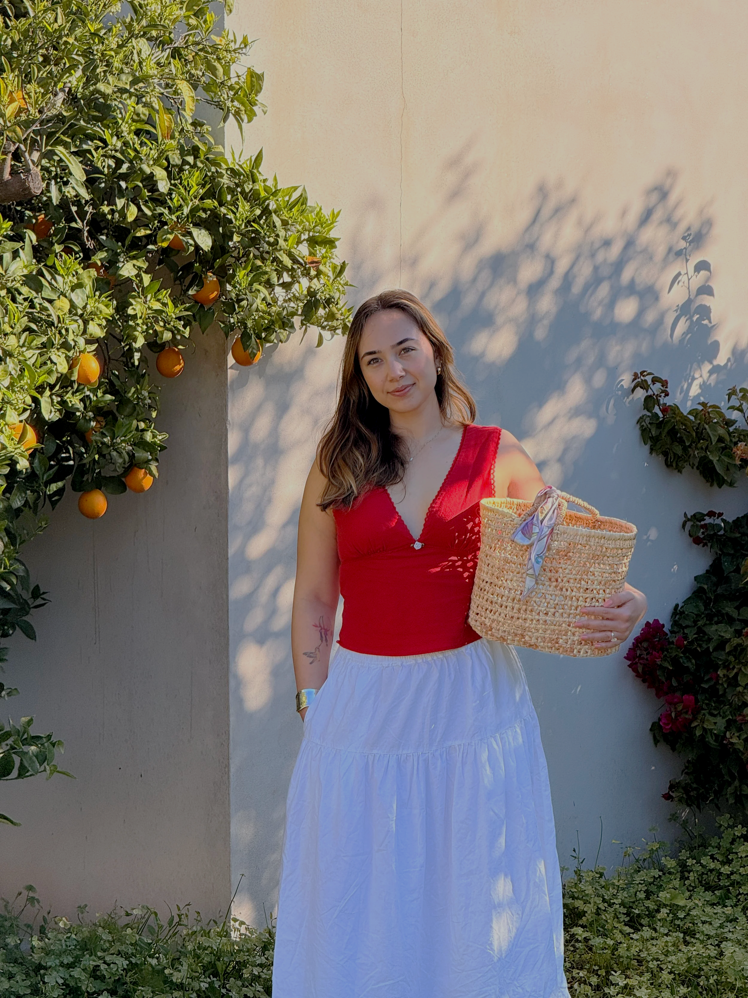 Founder of Solenne. a woman wearing a red sleeveless top and a long white skirt standing outdoors. She is holding a woven basket with a colorful scarf. Behind her are orange trees and flowering bushes, with sunlight casting shadows on a white wall.