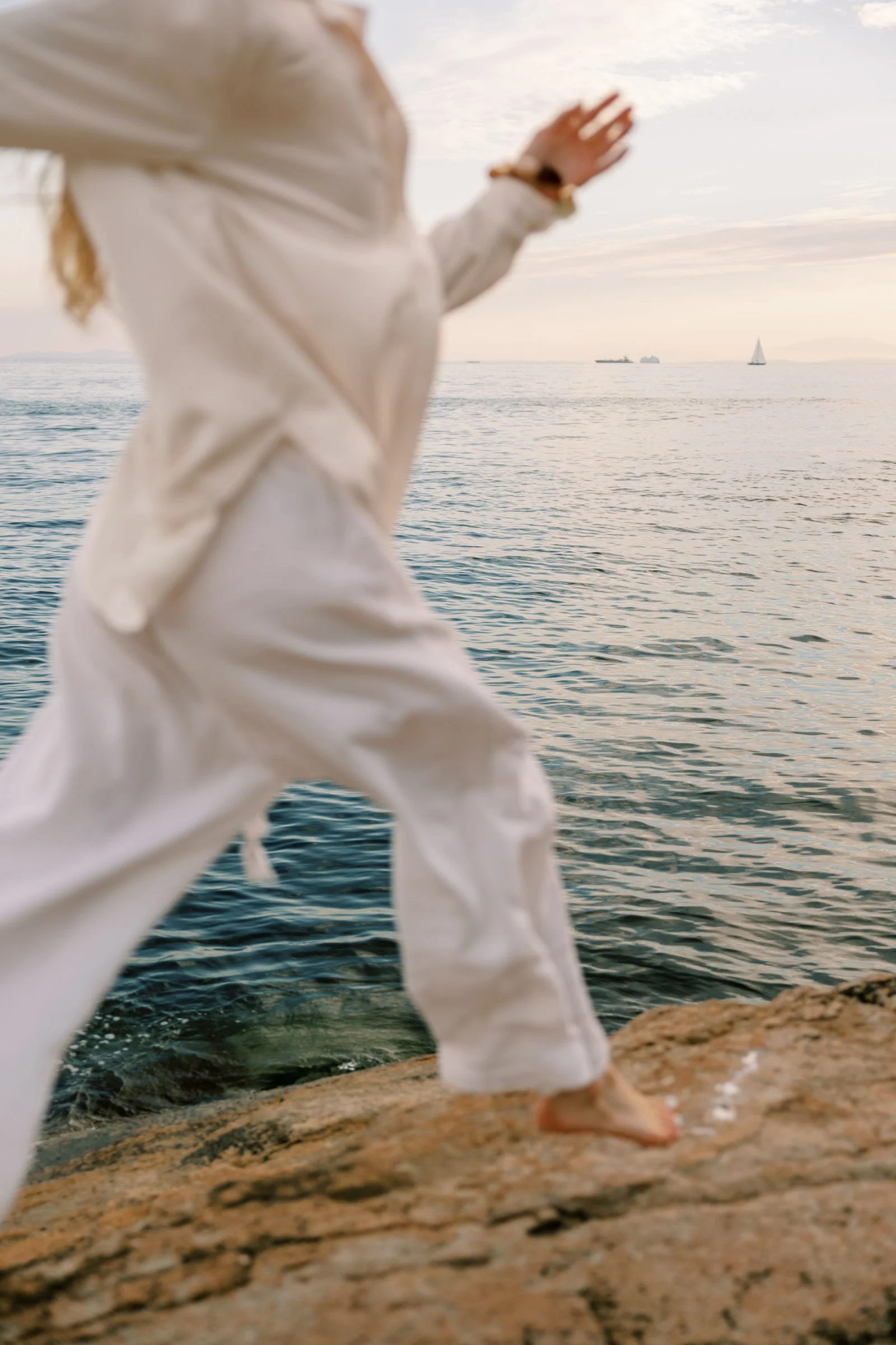 Woman standing on a rocky shoreline at sunset, warm coastal light and calm water — a scene from a women’s travel retreat by the sea.