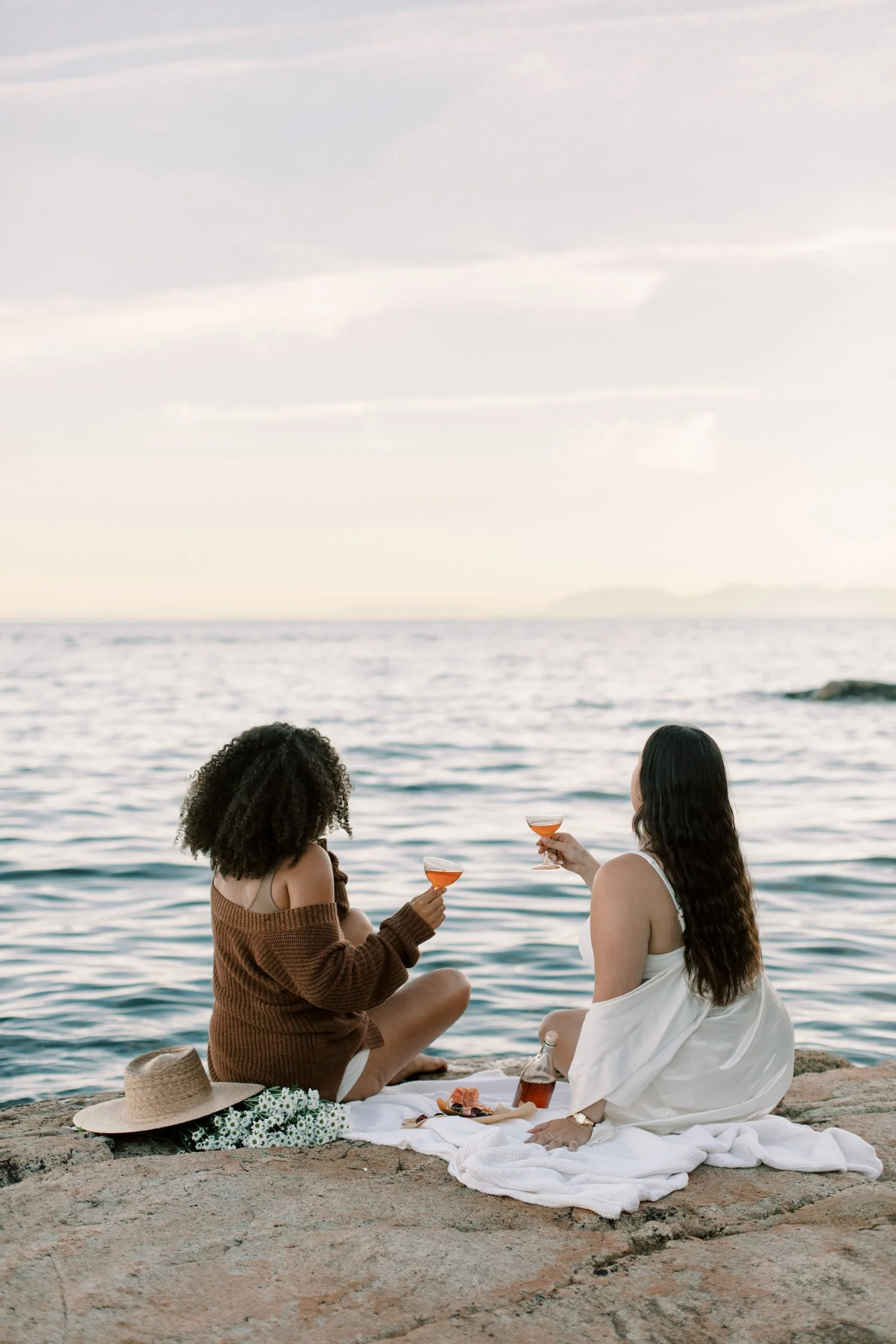 Women enjoying a sunset picnic on coastal rocks, toasting drinks by the water during a small‑group women’s travel retreat.