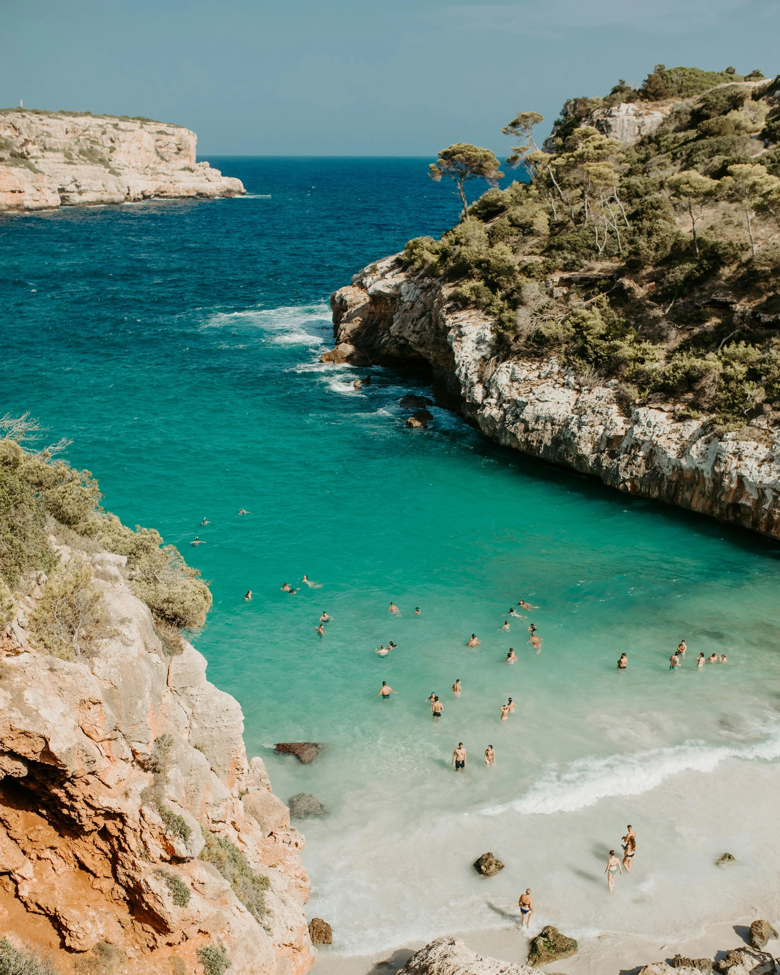 People swimming in a clear turquoise beach, surrounded by rocky cliffs and green shrubbery under a blue sky at Mallorca womens retreat