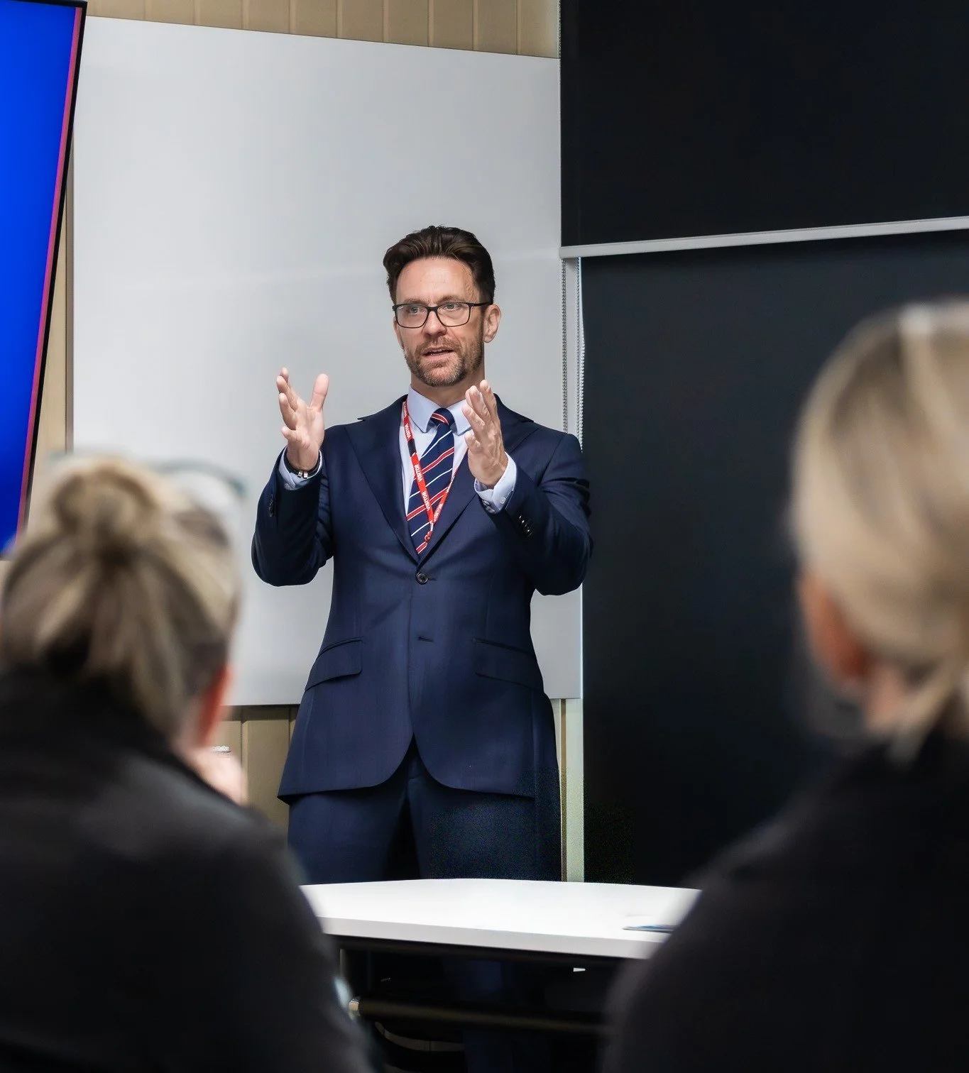 A man in a suit giving a presentation in front of an audience in a conference room.