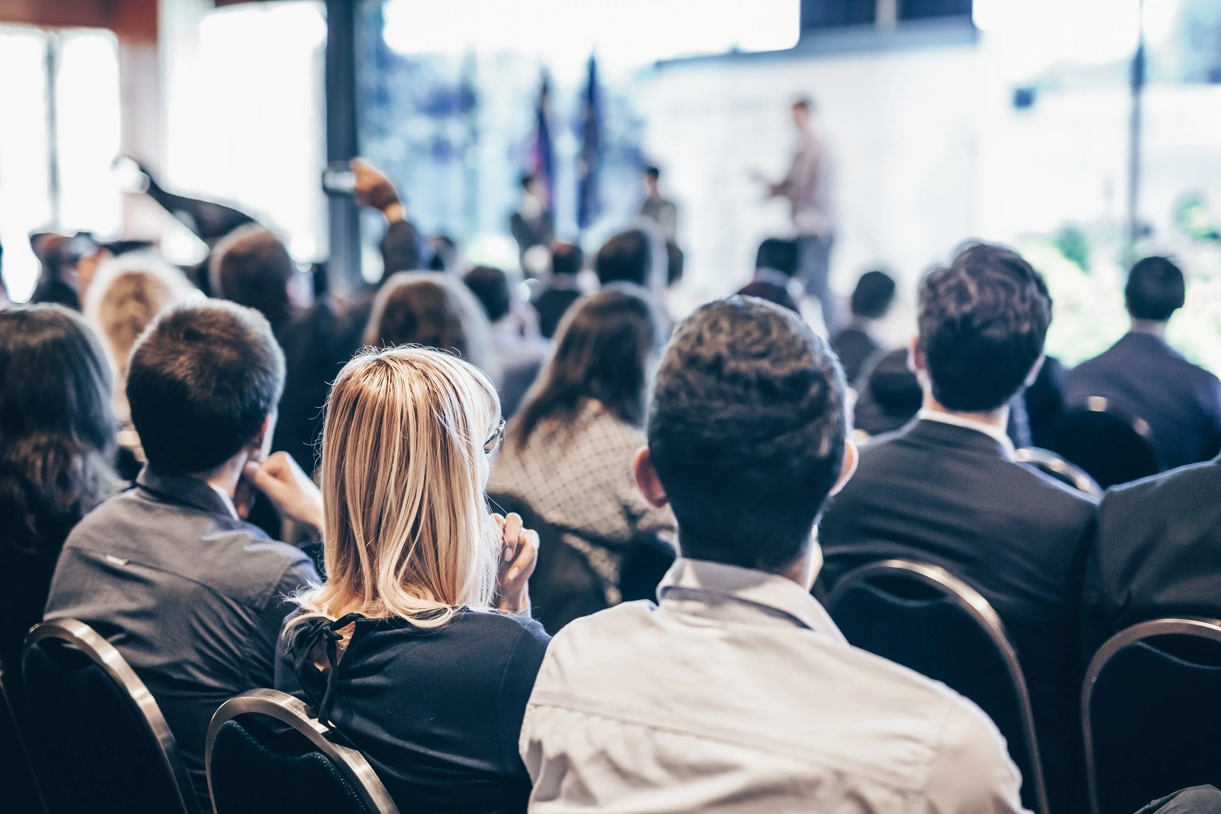 A large group of people attending a conference or seminar in a bright, modern venue, with some people taking notes or listening to a speaker at the front.