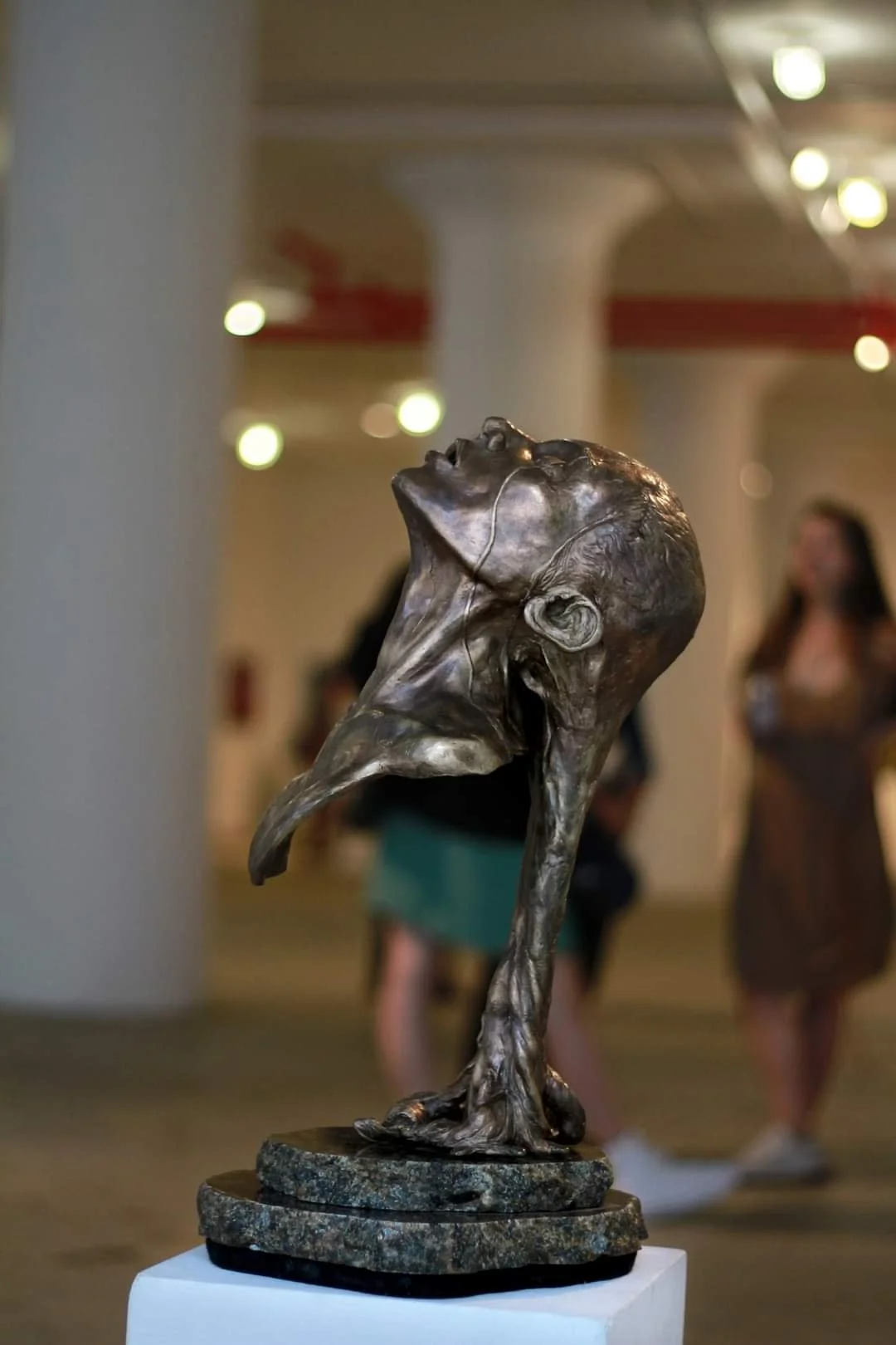 Bronze sculpture of a woman’s head tilted back, with short hair, mounted on a circular marble base, displayed in an art gallery with blurred people in the background.