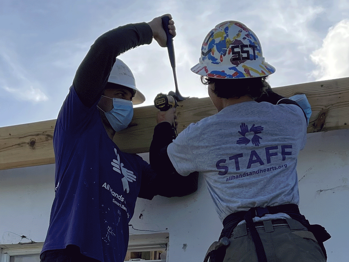 Two construction workers, wearing helmets and masks, are installing a wooden beam on a building's exterior wall.