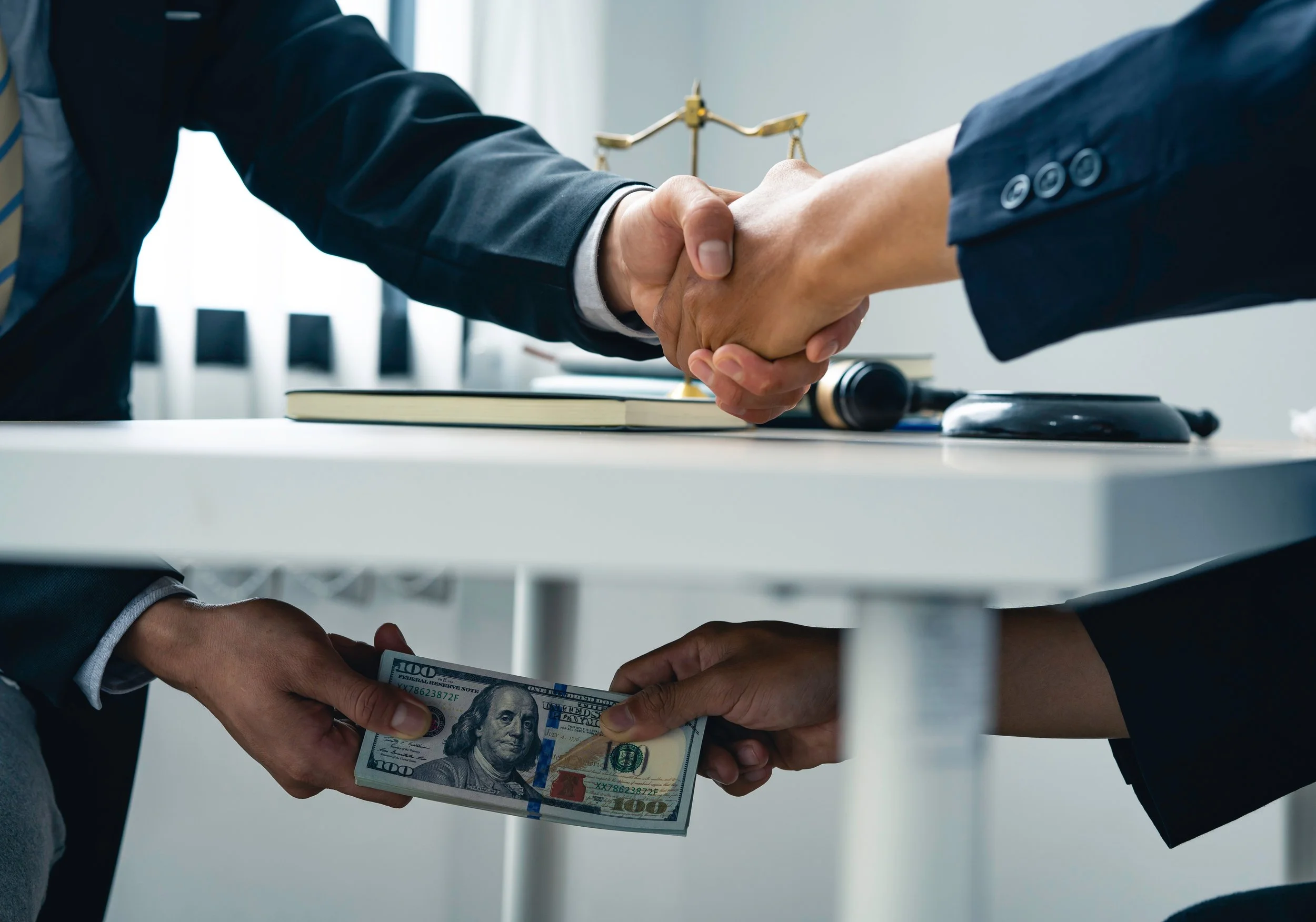 Two people shaking hands over a desk, with one person giving cash to the other beneath the desk.