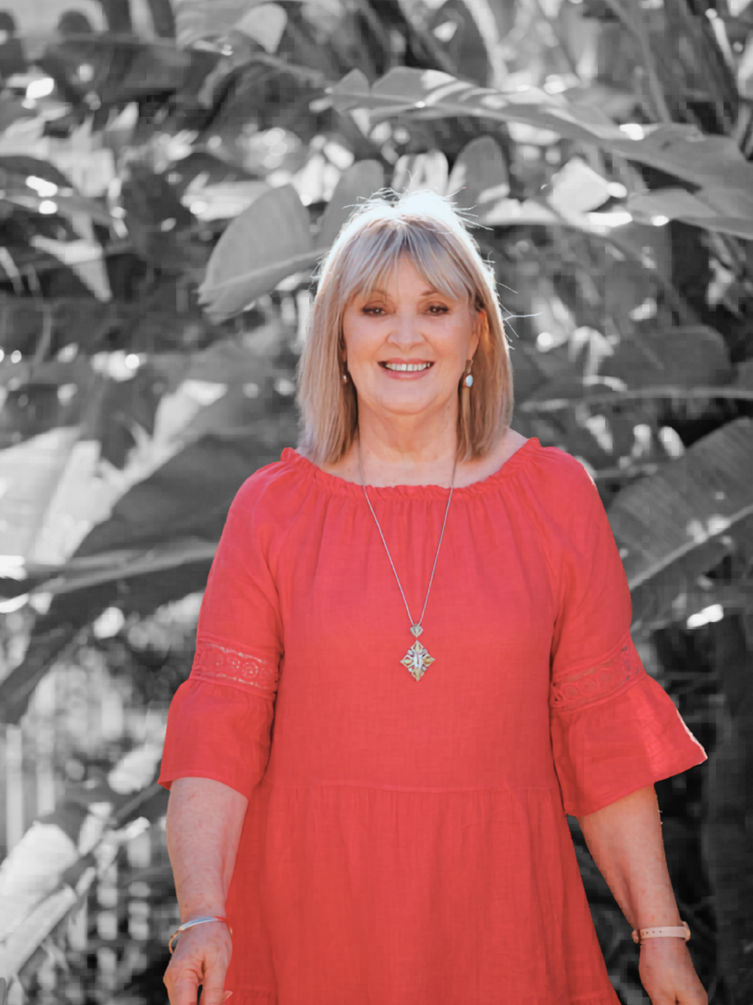 A woman with blonde hair wearing a red dress and earrings, smiling, standing in front of leafy plants