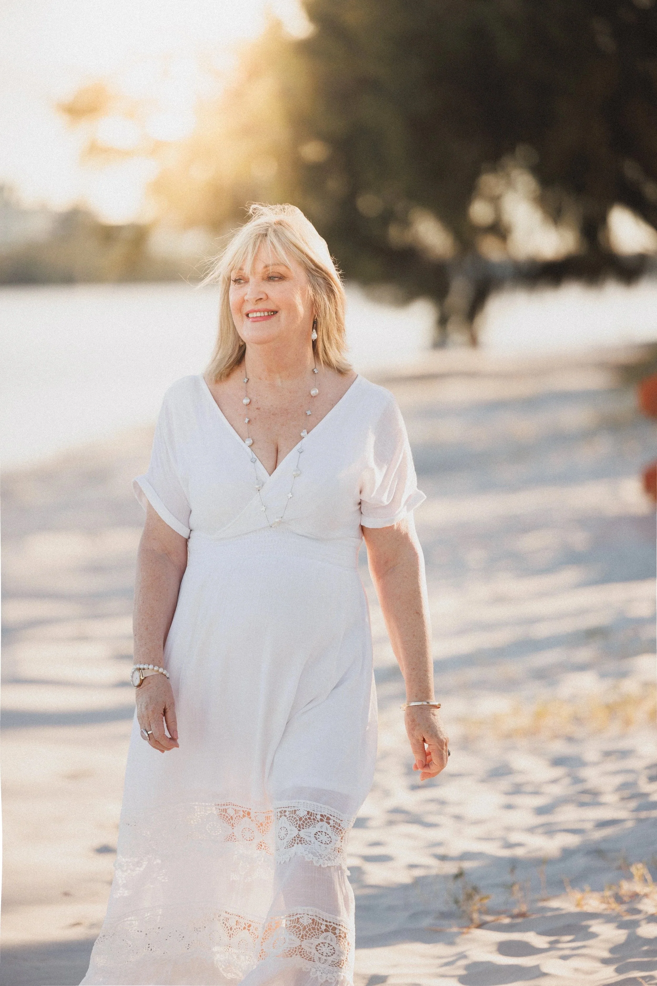A woman in a white dress standing on a sandy beach during sunset, smiling and looking to her left, with trees in the background.