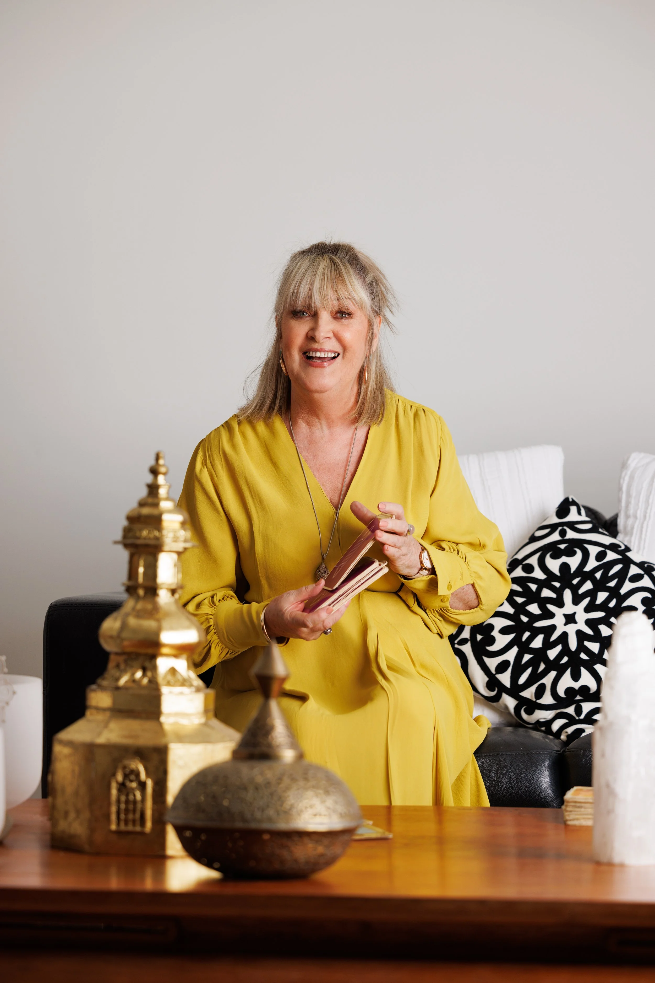 A woman with blonde hair and bangs wearing a yellow dress, sitting on a black couch, holding a notebook, smiling, with patterned pillows behind her. In the foreground, there are decorative items on a wooden table.